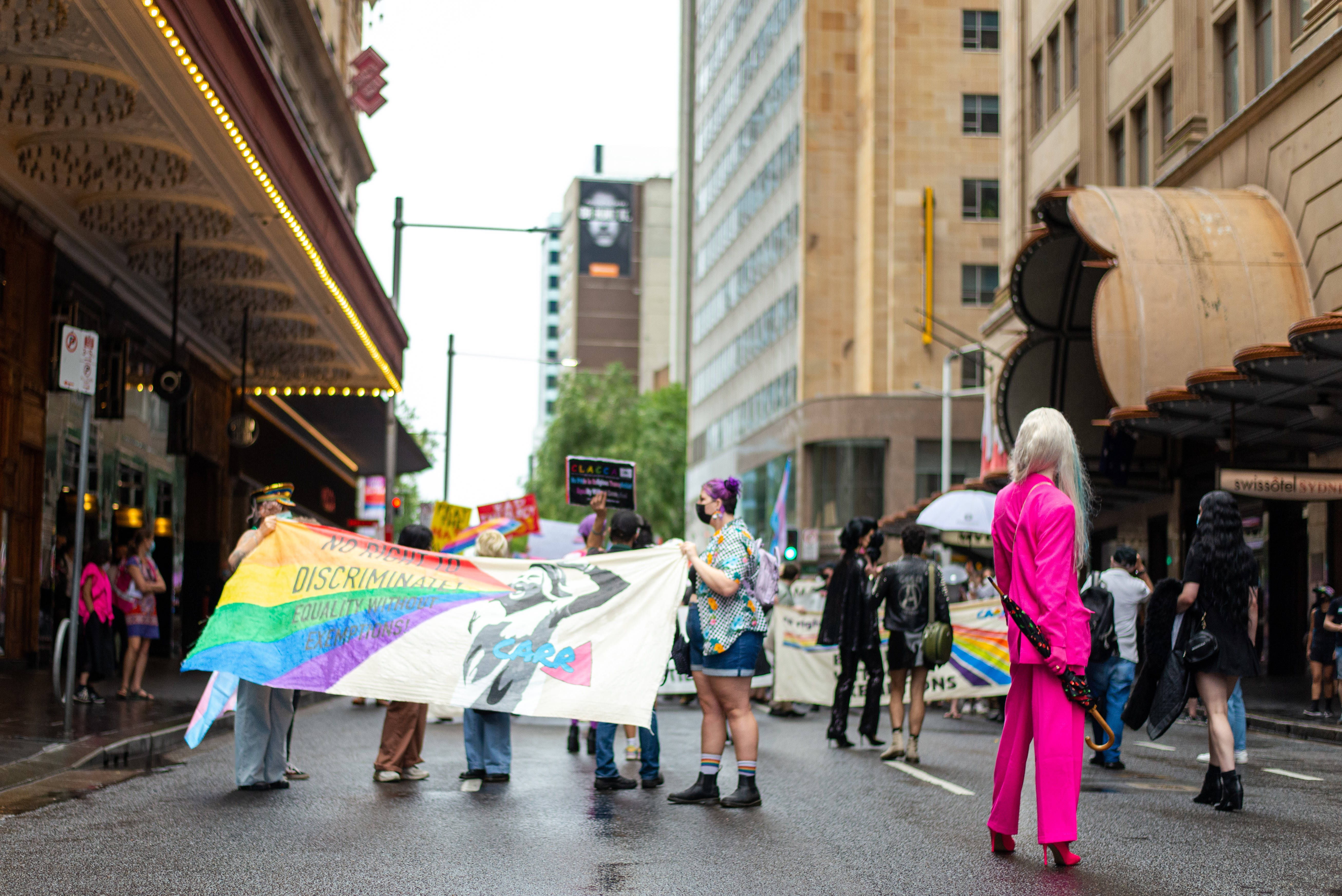 非二元驕傲旗幟迎風飄揚 壁紙 — 一場充滿活力的LGBTQ+驕傲遊行在濕漉漉的城市街道上展開，參與者高舉彩虹旗幟和標語，在多雲的天空下要求平等。