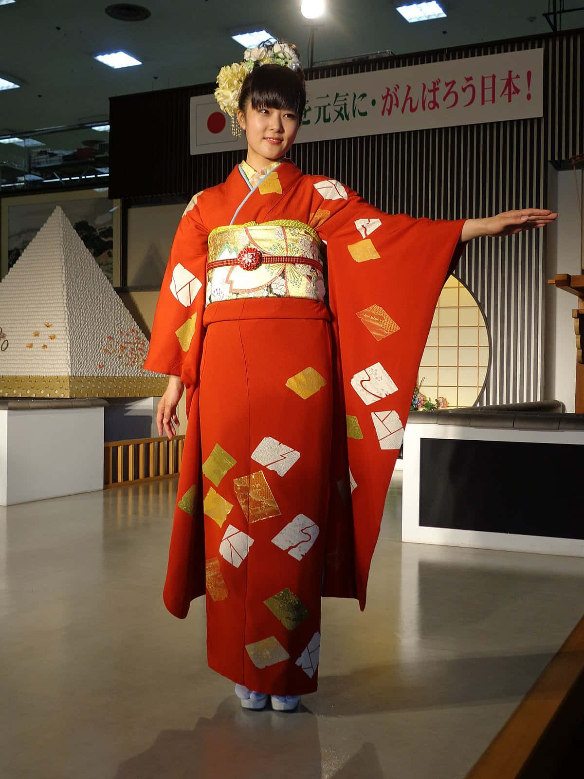 非凡的日本和服 壁紙 — 一位年輕女性身穿鮮紅色和服，帶有金色和白色的信封圖案，在明亮的燈光下優雅地在室內擺姿勢。
