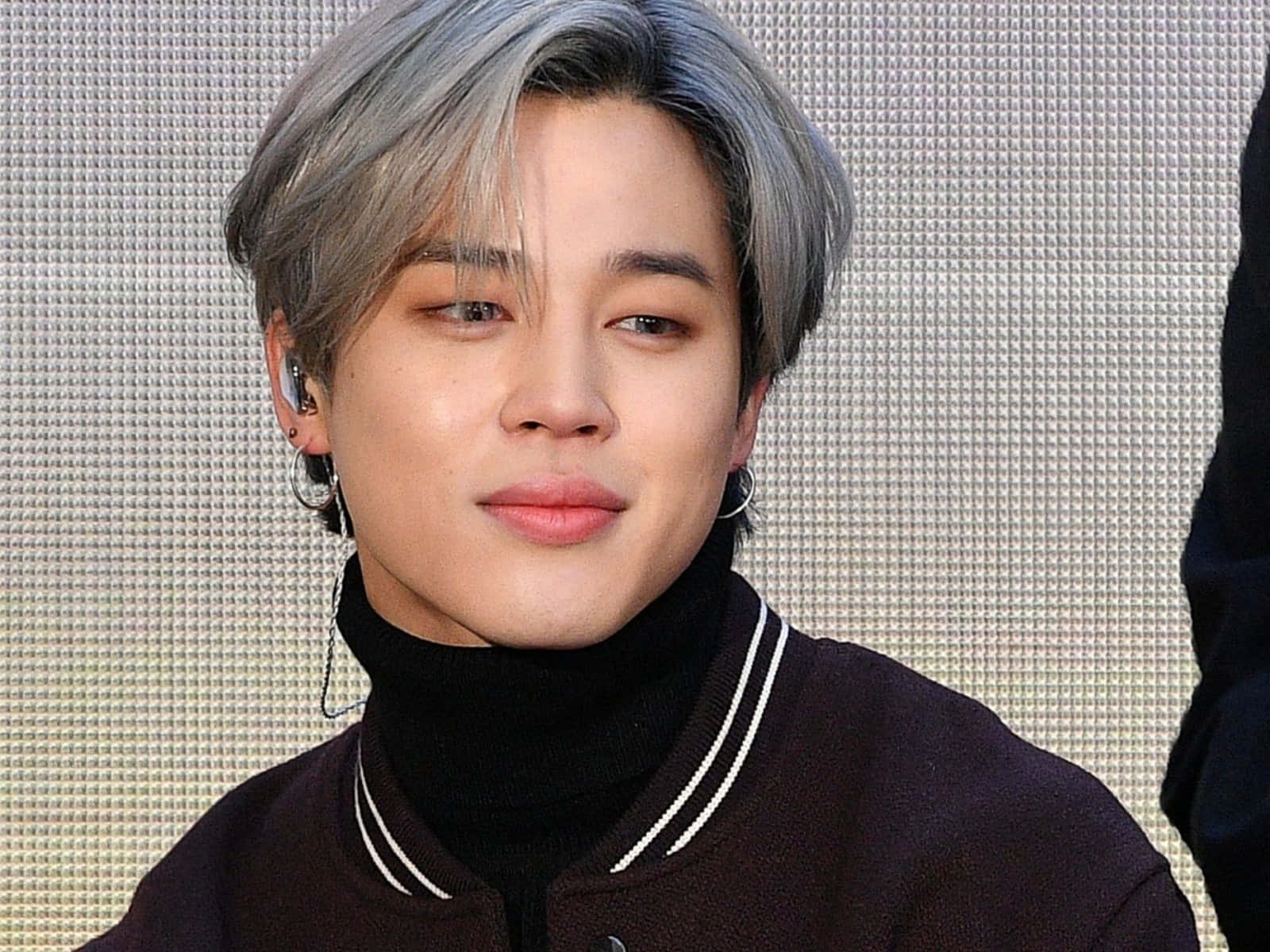 靠近音樂，感受快樂——與 Jimin 一同跳舞 壁紙 — 一位銀髮戴著耳環的年輕男子，穿著深色校隊夾克，背景是紋理牆。