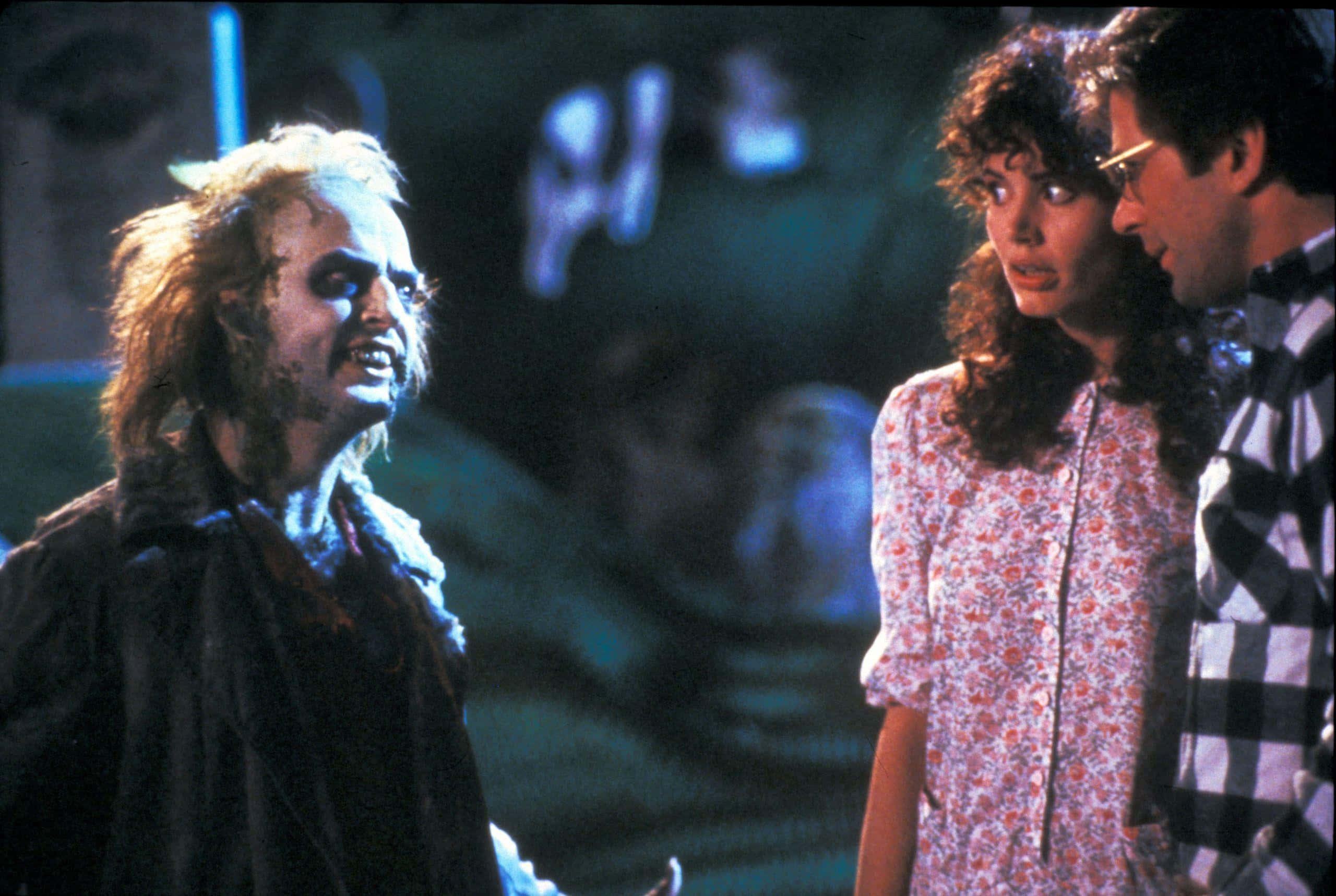 Beetlejuice 桌布 壁紙 — 面色蒼白、頭髮凌亂的Beetlejuice在昏暗的藍色燈光下與一位身穿碎花裙的女人和一位身穿格仔恤衫的男人對峙。