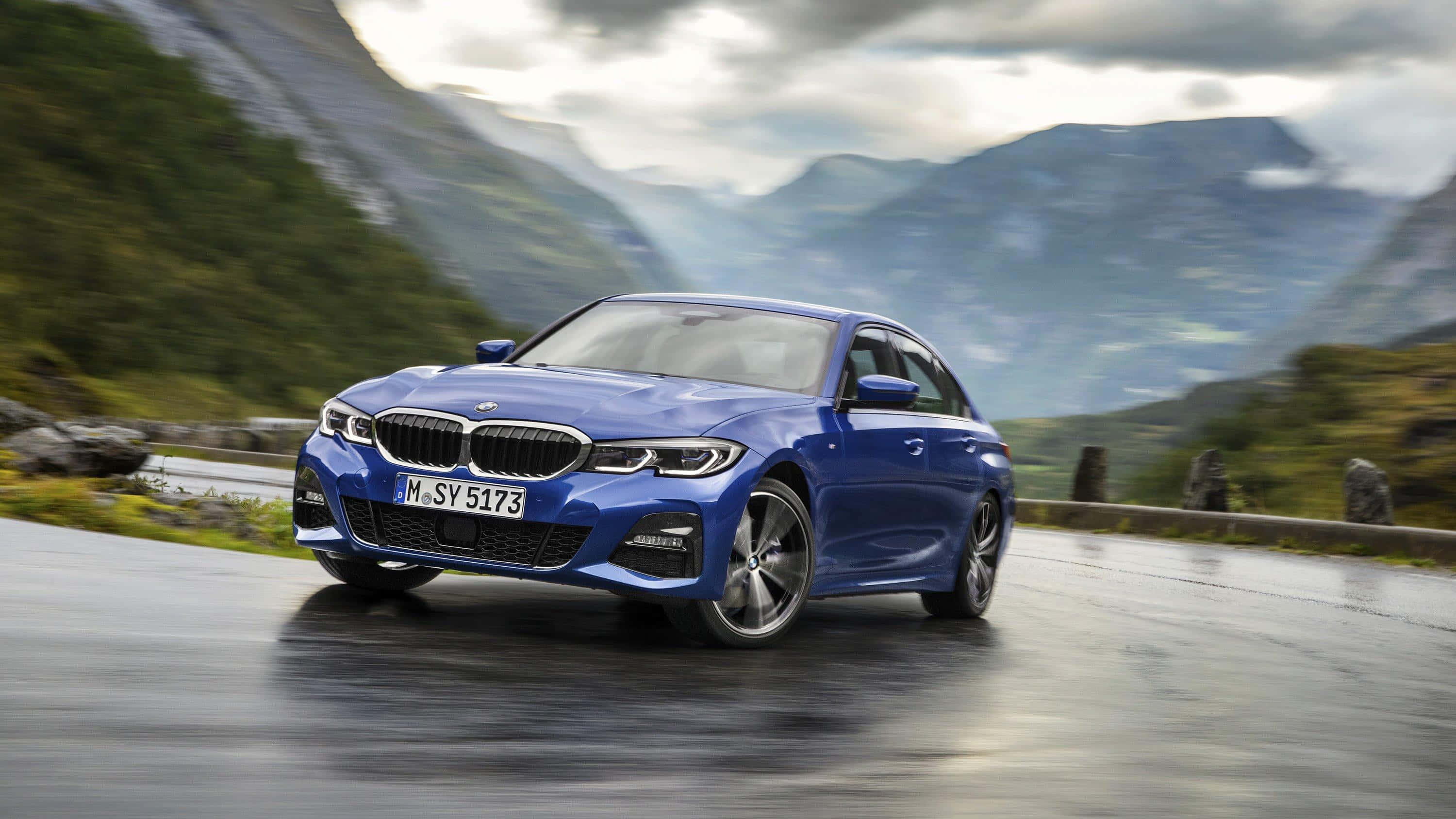 革新移動體驗：推出 BMW M 系列 壁紙 — 一輛充滿活力的藍色BMW房車在潮濕的山路上疾馳，兩旁是茂密的綠色山坡和霧氣繚繞的山峰，天空多雲。
