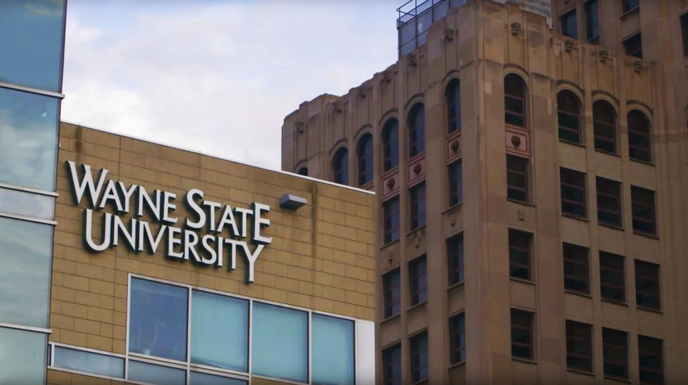 韋恩州立大學校園入口標誌 壁紙 — Wayne State University 建築物，擁有現代玻璃外牆和經典石塔，在陰天天空下。