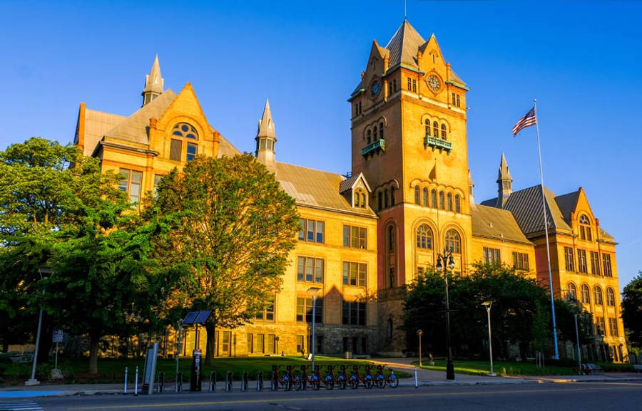 韋恩州立大學校園風景 壁紙 — 一座宏偉的歷史石造法院大樓沐浴在金黃色的日落光芒中，周圍有茂密的綠樹和飄揚的美國國旗。
