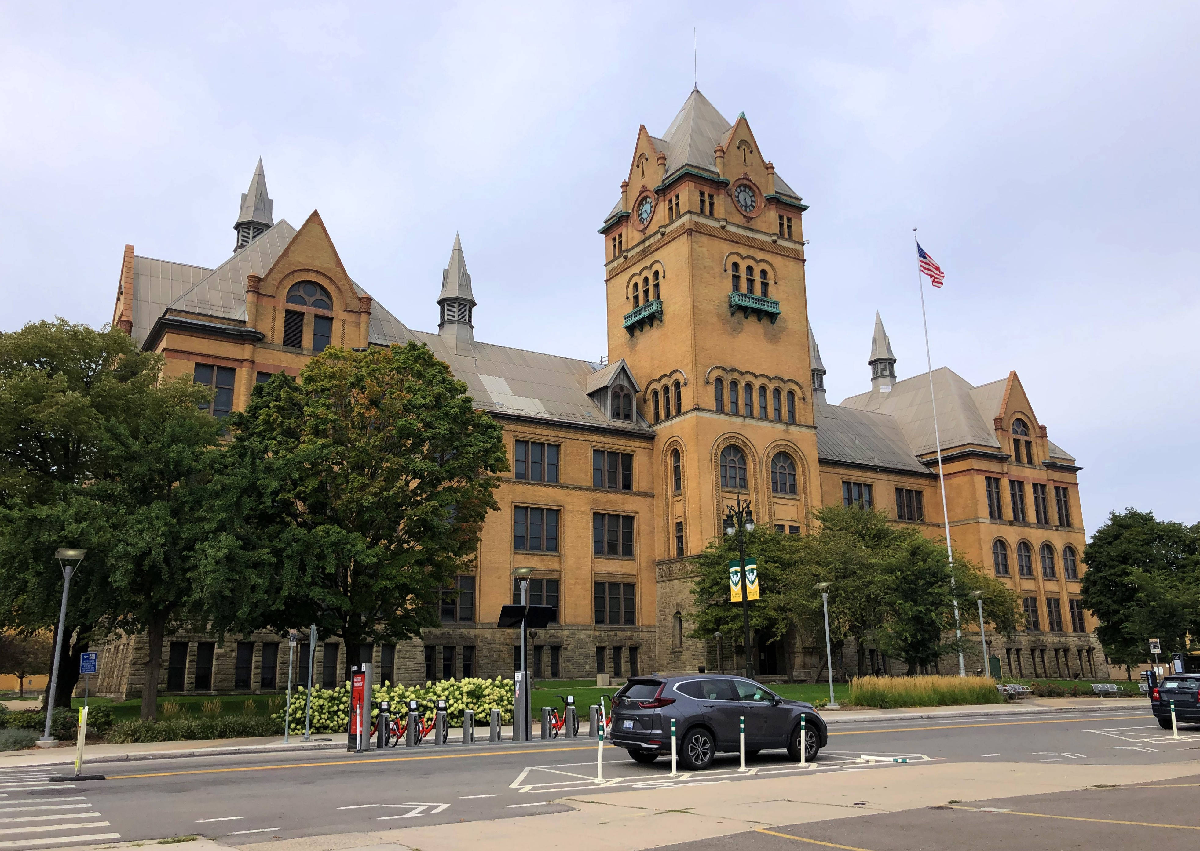 韋恩州立大學舊主樓與天空背景 壁紙 — 一座宏偉的歷史砂岩建築，帶有鐘樓和美國國旗，矗立在陰天之下，前面有樹木和一輛停著的汽車。