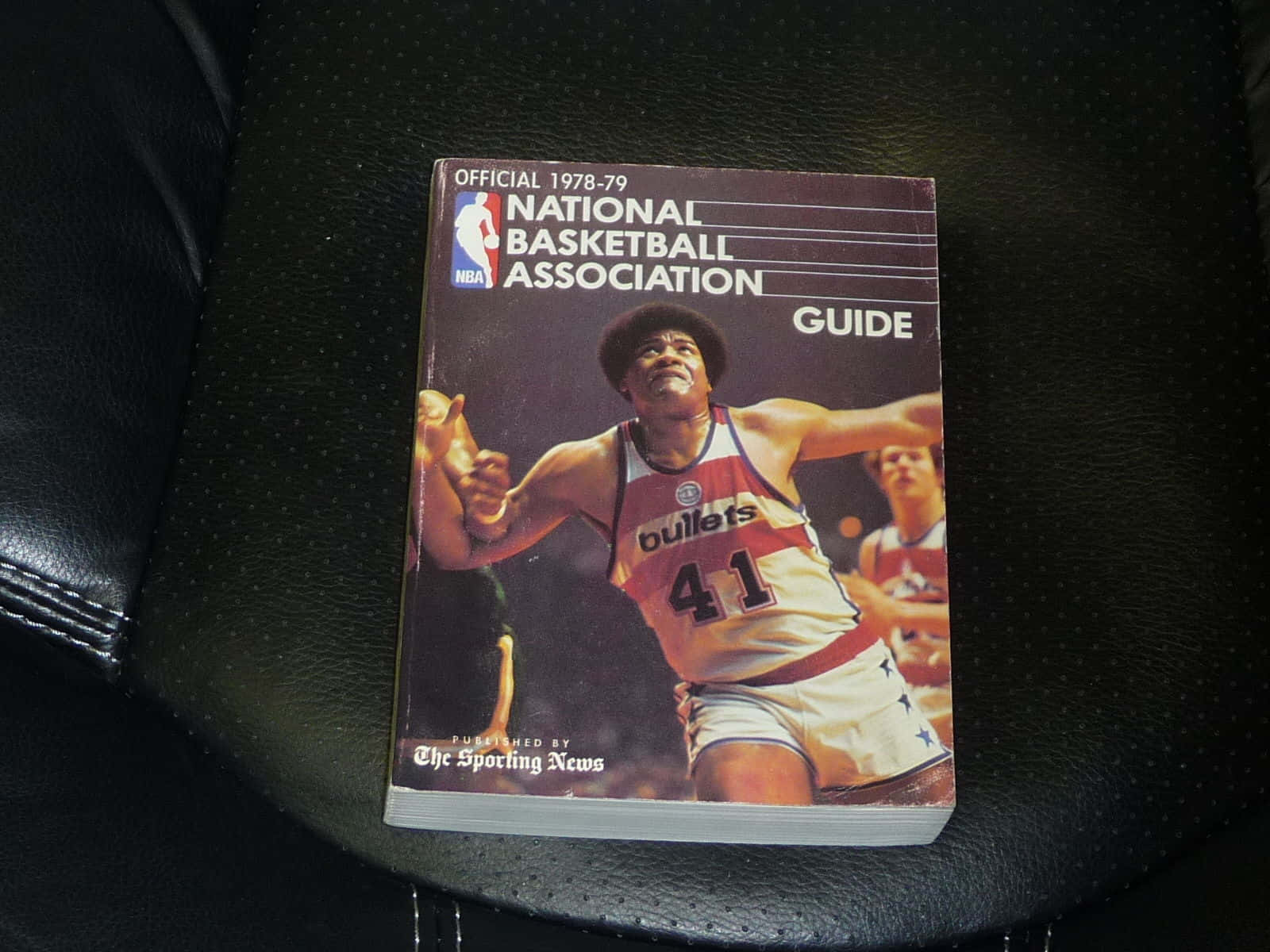 韋斯·安斯利封面NBA指南 壁紙 — 一本1978-79年復古NBA指南，上面有穿著子彈隊球衣的籃球運動員，放在黑色皮革座椅上。