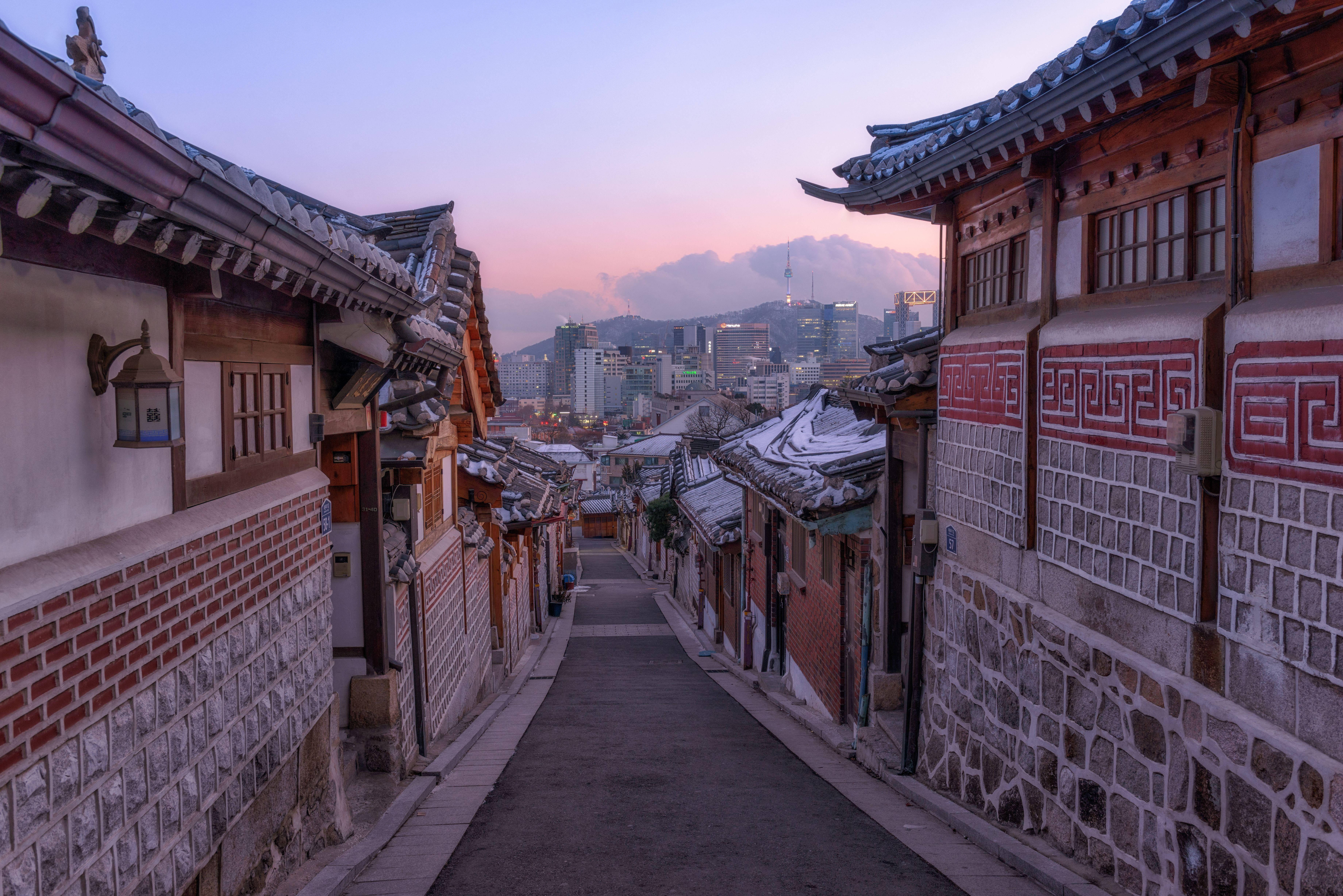 韓國北村韓屋村 壁紙 — 一條狹窄的傳統韓國小巷，兩旁是石頭和木屋，通向黃昏時分的現代城市天際線。