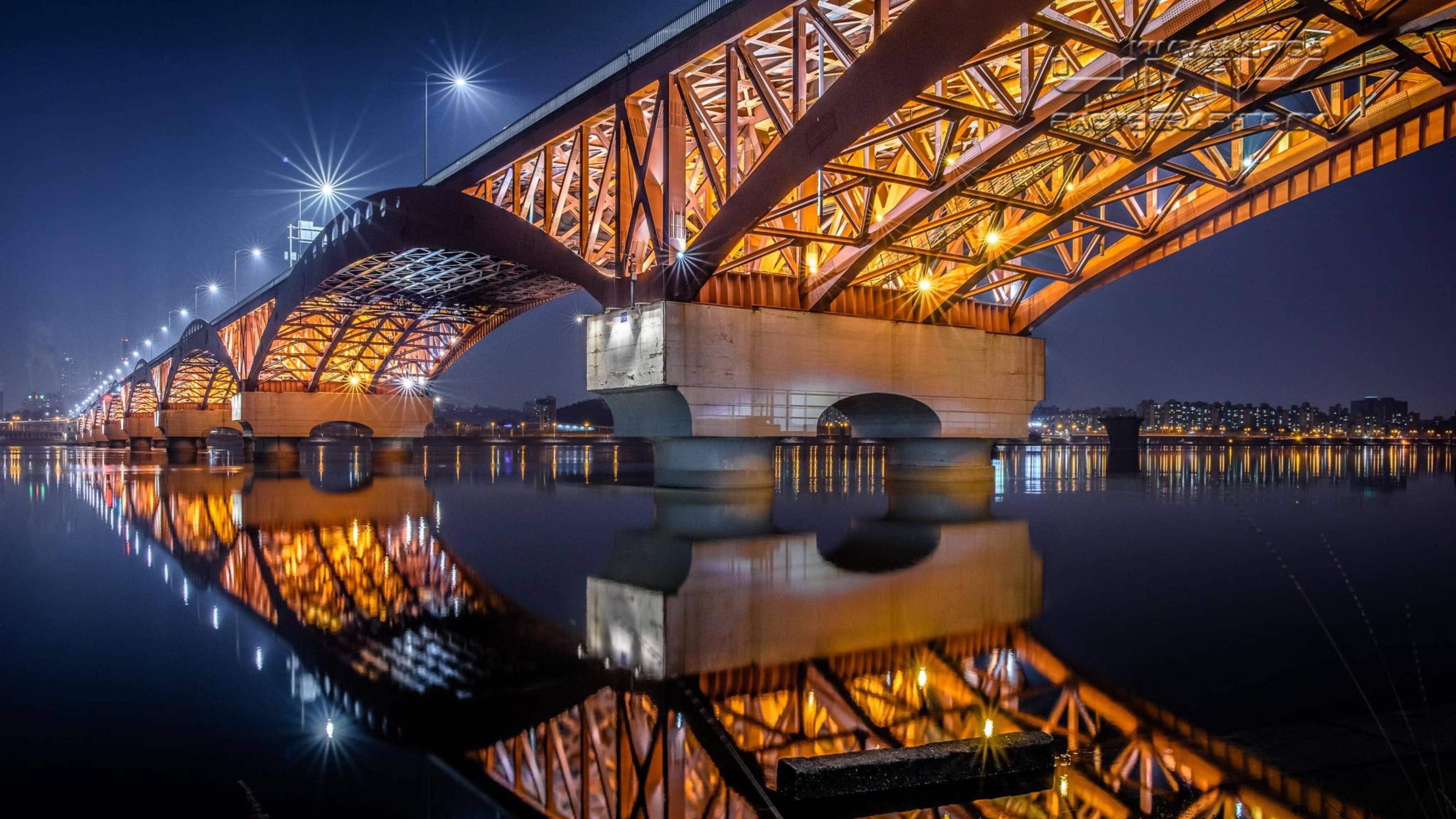 韓國城市夜景 壁紙 — 一座宏偉的照明鋼橋在夜間拱跨平靜的水面，其溫暖的燈光營造出完美的鏡面倒影。
