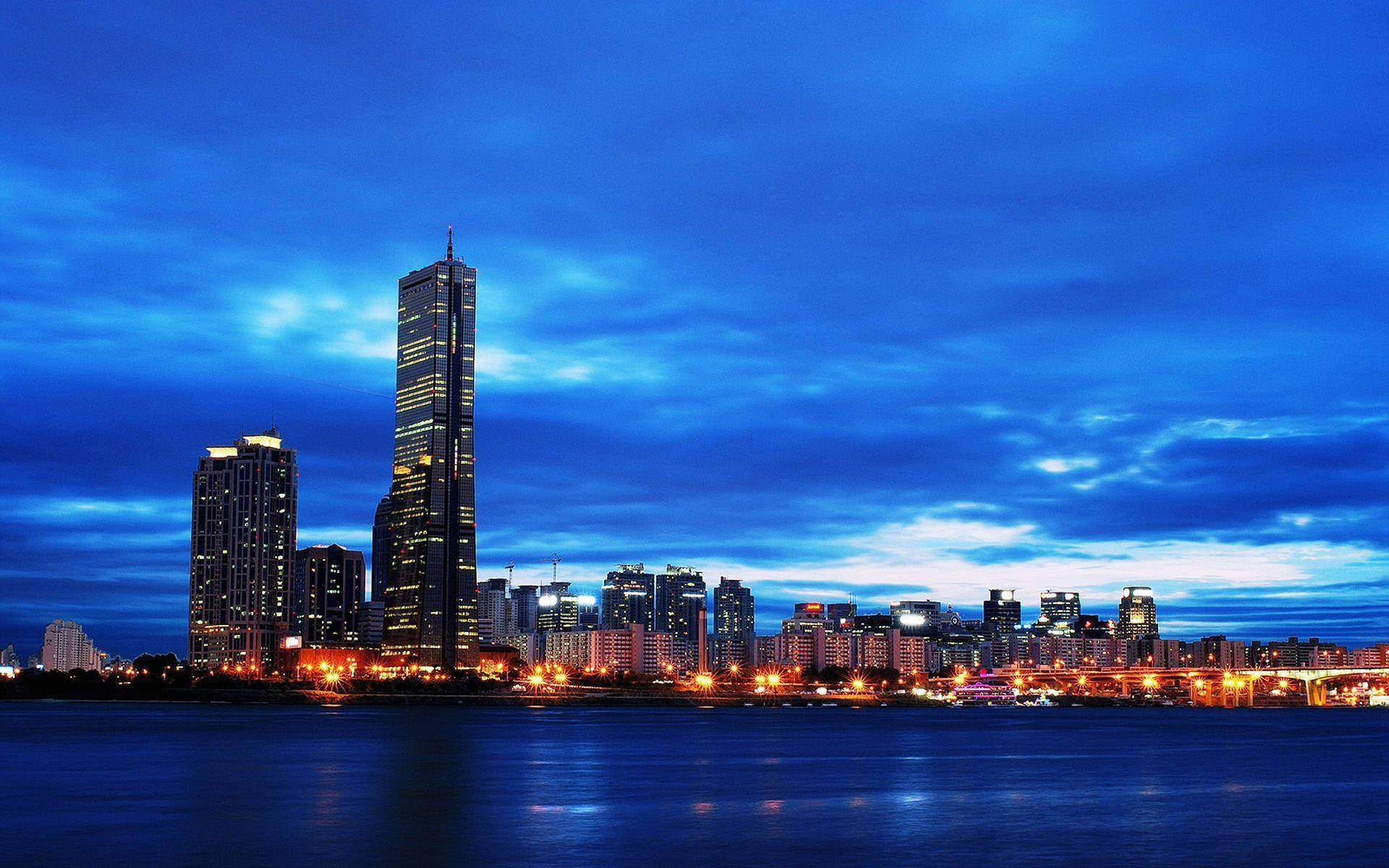韓國城市天際線 壁紙 — 令人驚嘆的黃昏城市景觀，高聳的摩天大樓在深藍色天空下，將溫暖的燈光反射在平靜的水面上。