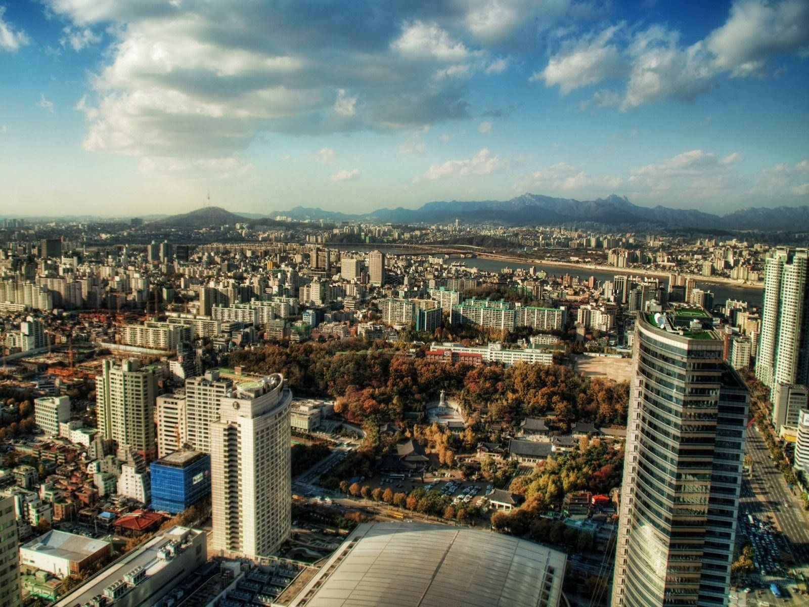 韓國天空 壁紙 — 俯瞰廣闊的城市景觀，秋樹與遠山在部分多雲的天空下。