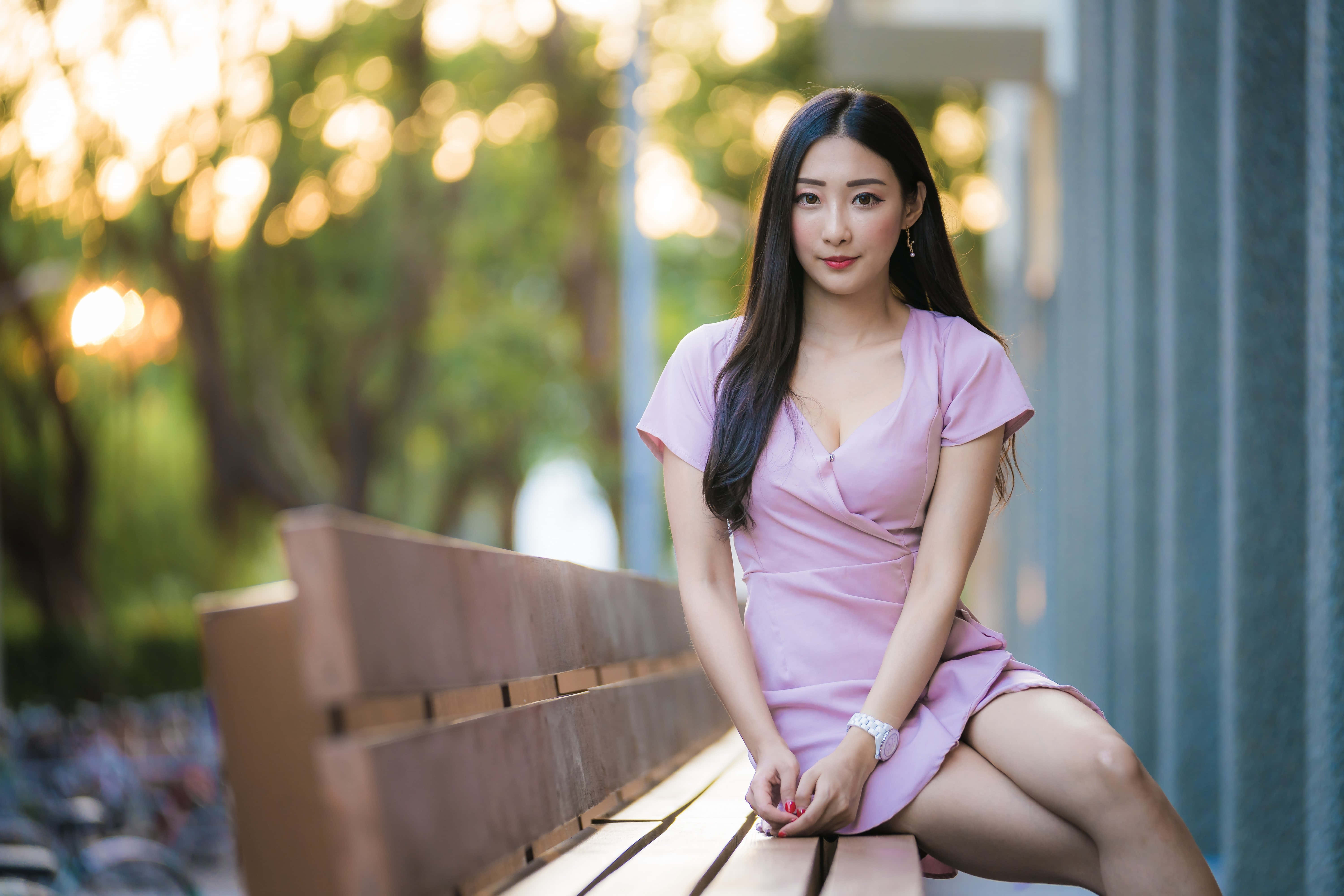 韓國女性穿紫色高衩裙坐長凳 壁紙 — 一位身穿粉紅色連衣裙的女性優雅地坐在木長椅上，金色陽光透過她身後的樹木灑落。