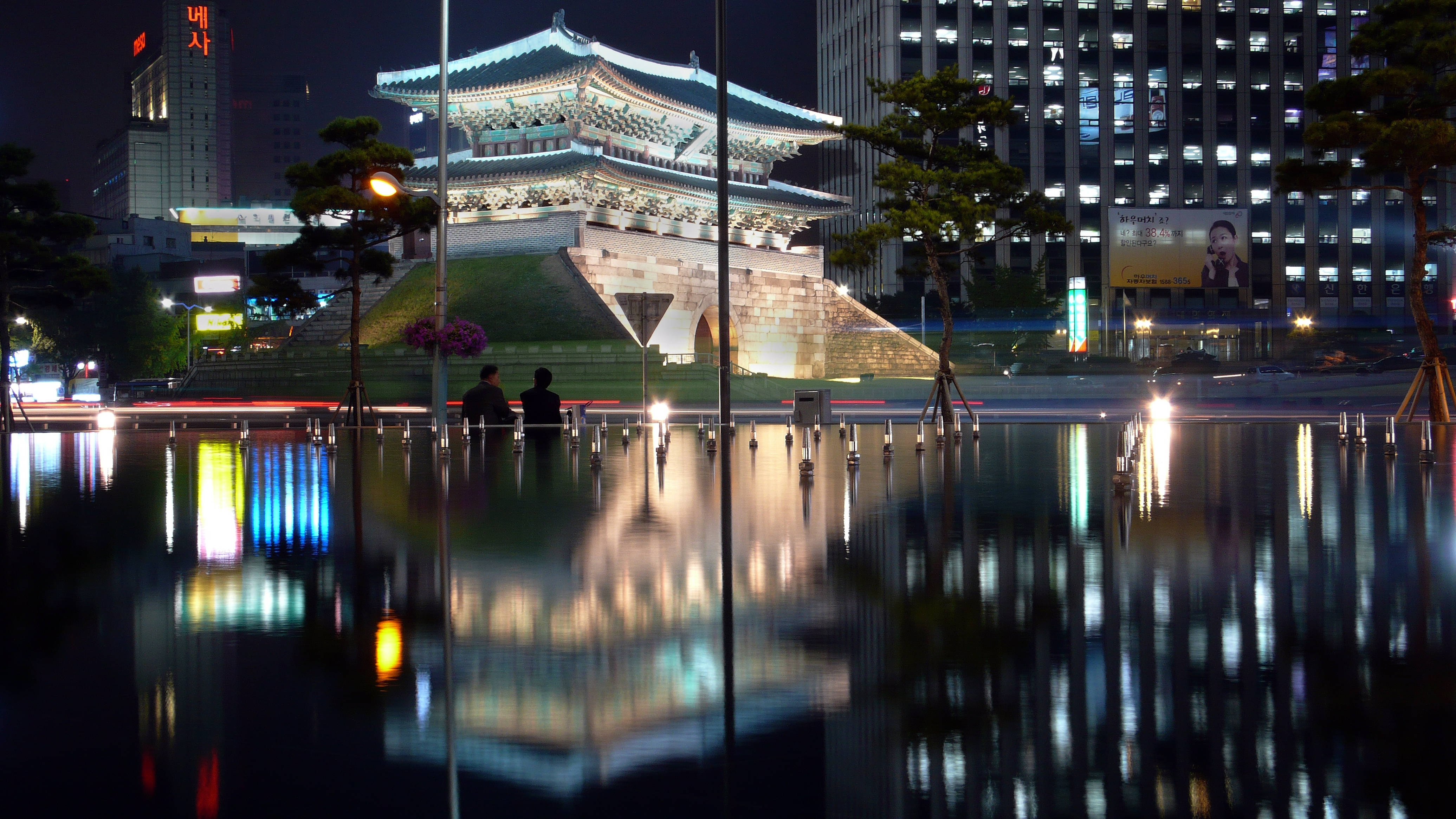 韓國成均館門 壁紙 — 一座歷史悠久的韓國城門在夜晚發光，燈光倒映在平靜的水池中，背景是模糊的城市燈光。