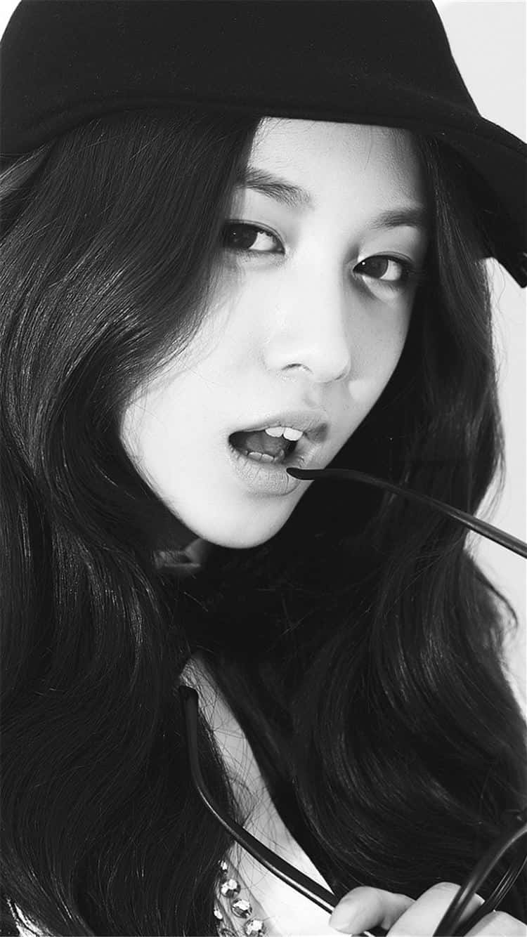韓國歌手朴志妍黑白肖像照 壁紙 — 一位長黑髮女性戴著帽子，手持太陽眼鏡靠近嘴巴的黑白肖像。