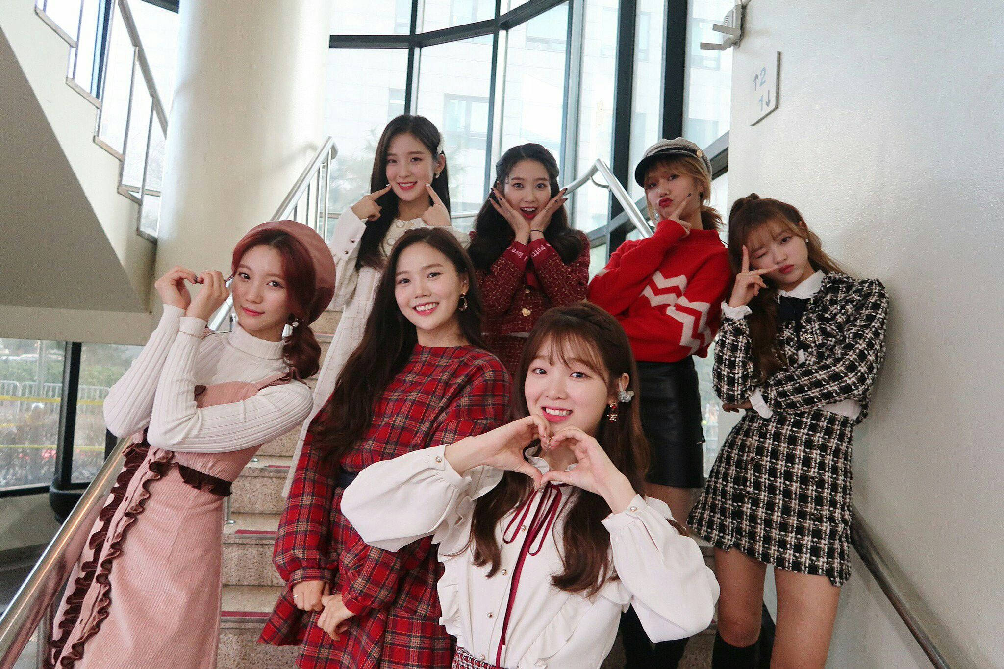 韓國流行天團OH MY GIRL 真人合照 壁紙 — 七位時尚年輕女性在現代室內樓梯上愉快地擺姿勢，身後有大型窗戶。