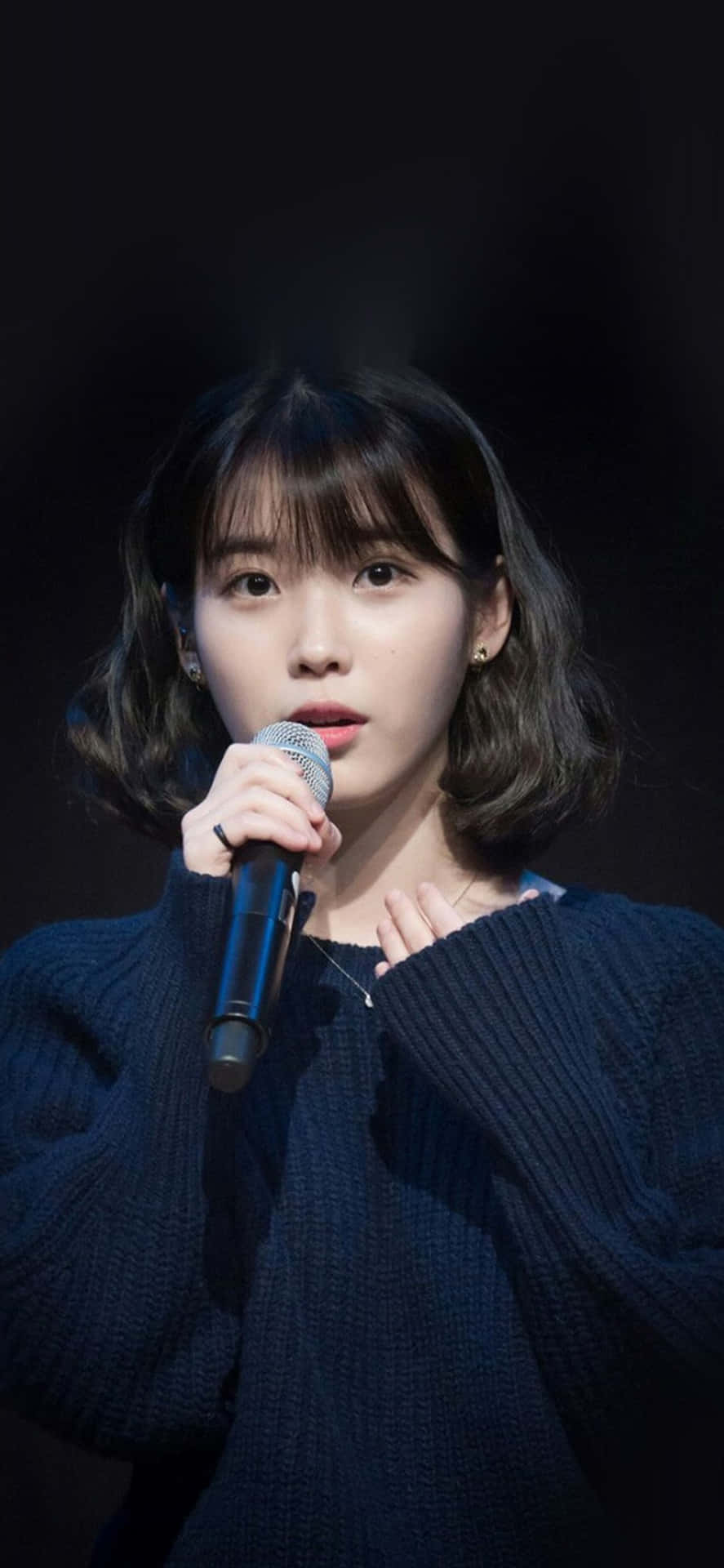韓國流行歌手展現舞台魅力 壁紙 — 一位短黑髮的年輕女子對著麥克風唱歌，身穿深藍色針織毛衣，背景為黑色。