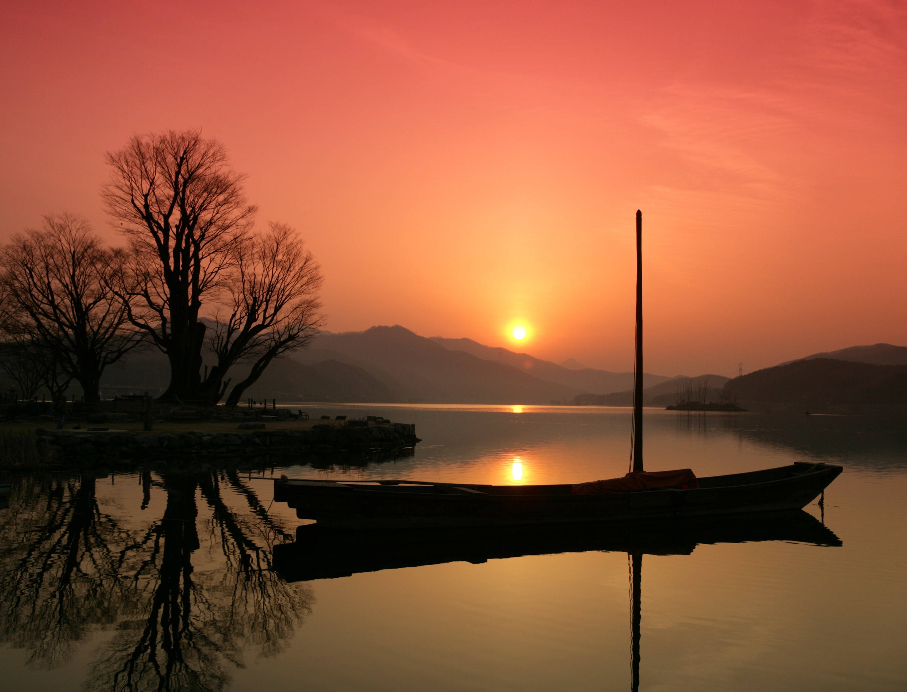 韓國湖畔夕照 壁紙 — 一艘孤獨的小船在日落時分停泊在平靜的水面上，光禿禿的樹木和遠山在火紅的橙色天空下形成剪影。
