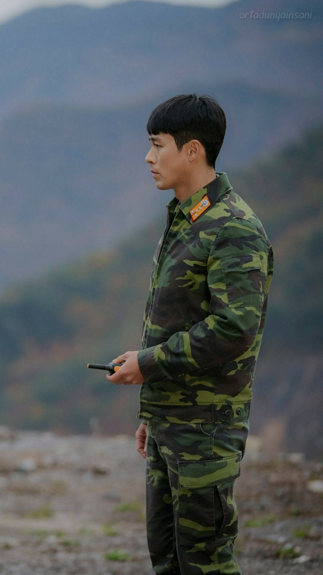 韓國演員玄彬 壁紙 — 一名身穿迷彩軍服的士兵獨自站在岩石山坡上，俯瞰霧氣繚繞的山巒，手持無線電。