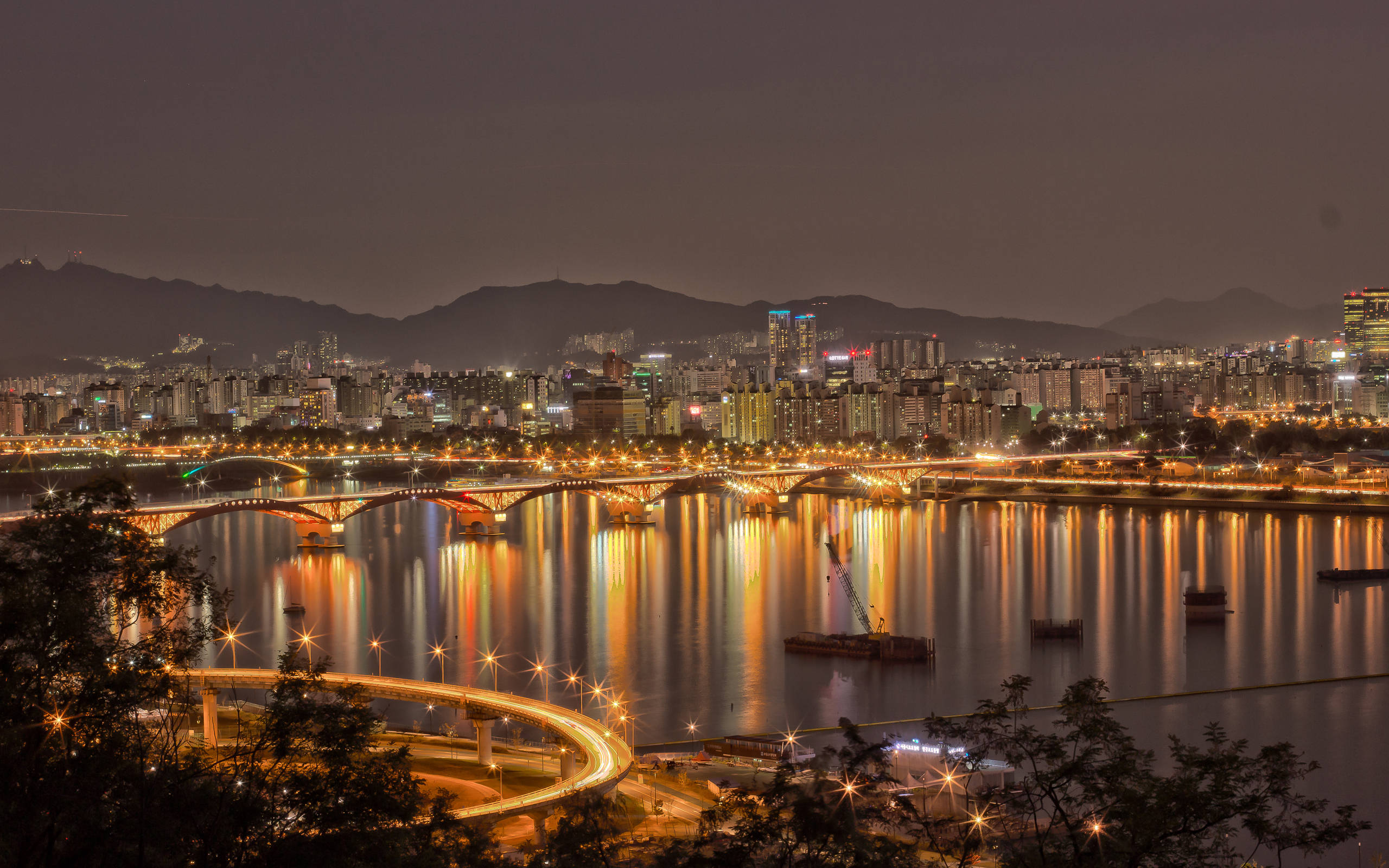 韓國漢江夕照 壁紙 — 充滿活力的夜景，發光的橋樑和摩天大樓倒映在平靜的河流中，周圍環繞著黑暗的山脈。