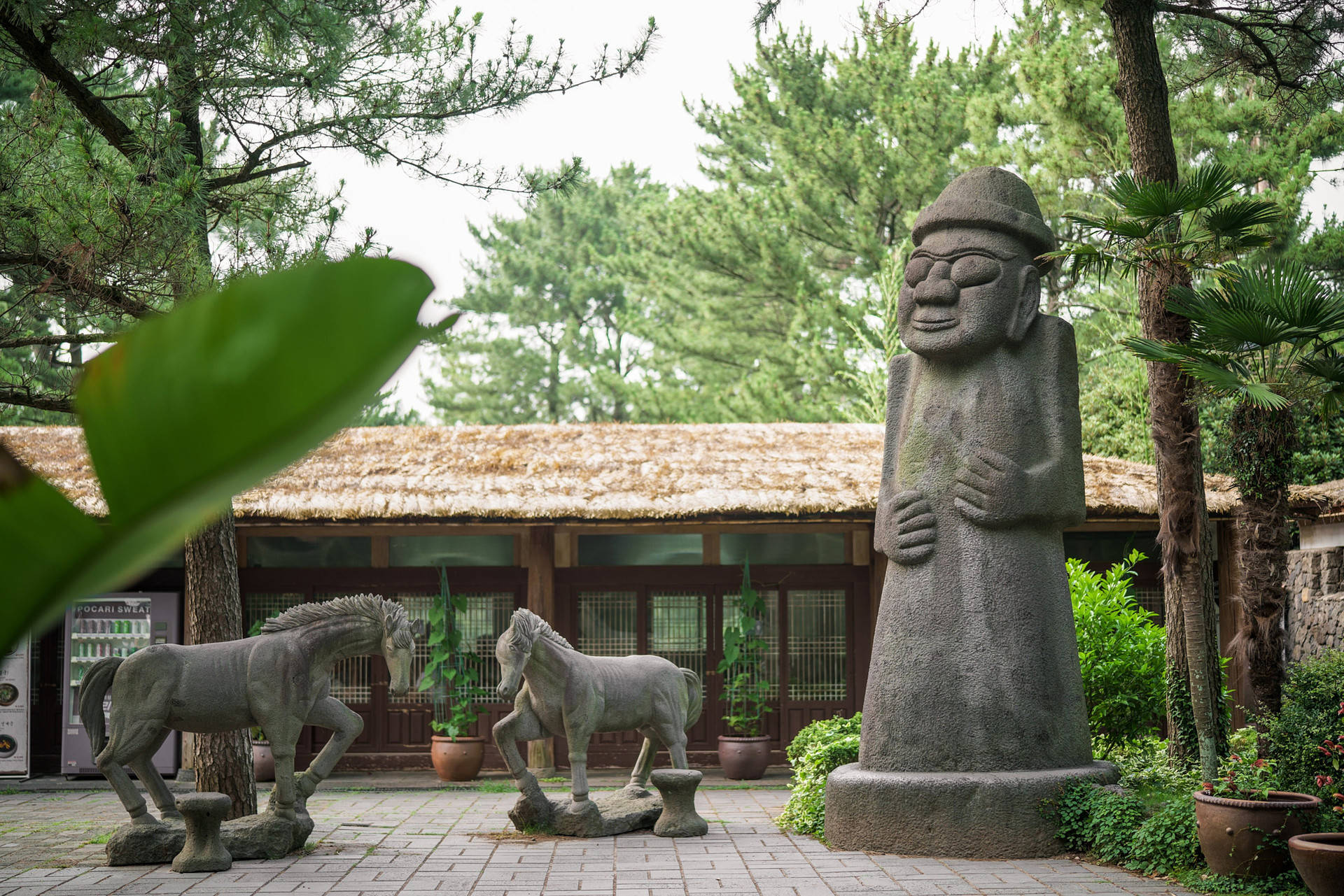 韓國濟州島民俗村風景 壁紙 — 一個寧靜的庭院中，有一座微笑人物的高聳石像，旁邊有兩匹馬的雕塑，周圍是茂密的綠樹。