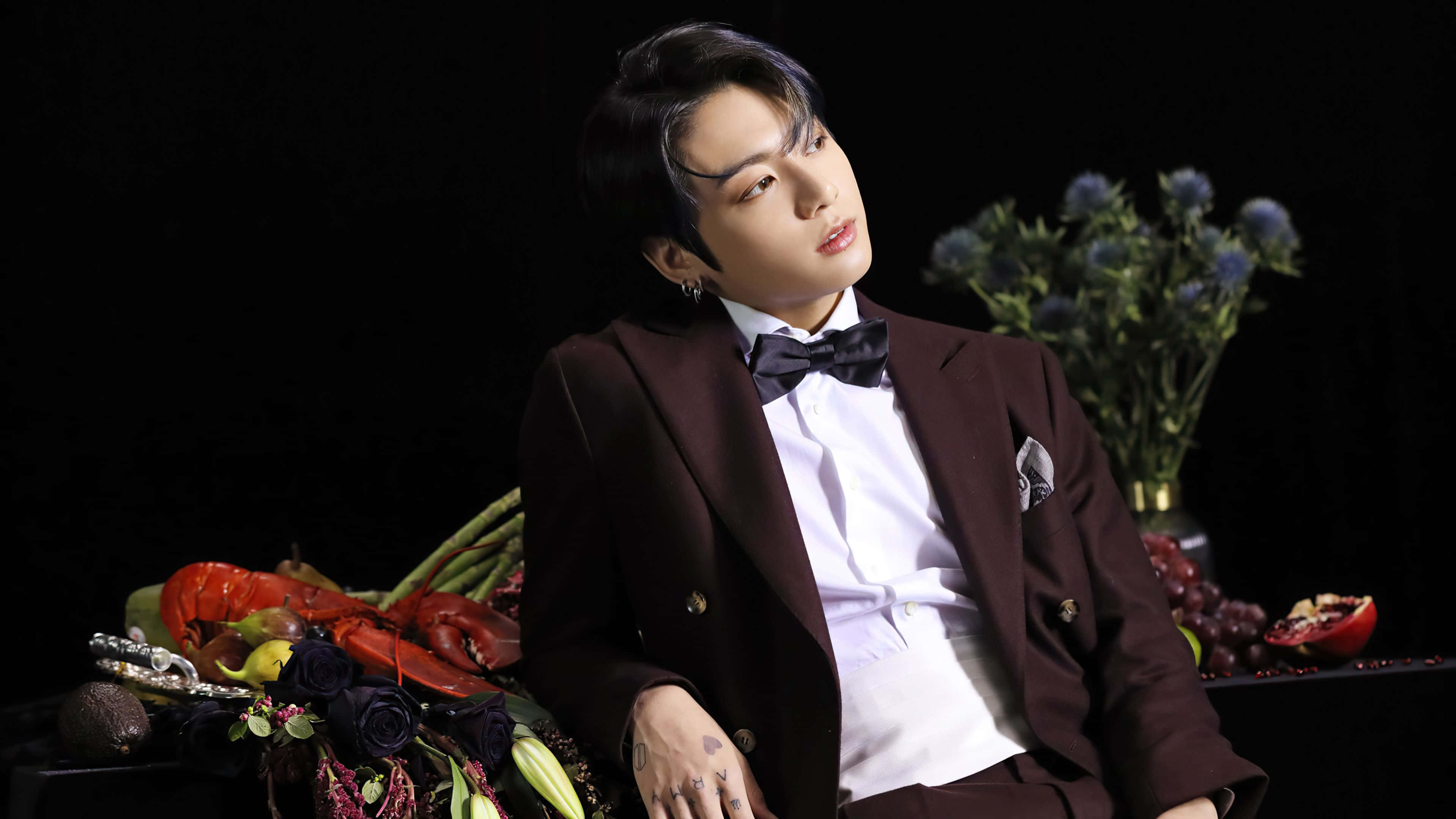韓國男子團體BTS成員Jungkook 壁紙 — 一名身穿栗色西裝和領結的男子在黑色背景前優雅地擺姿勢，周圍環繞著鮮豔的花朵和水果。