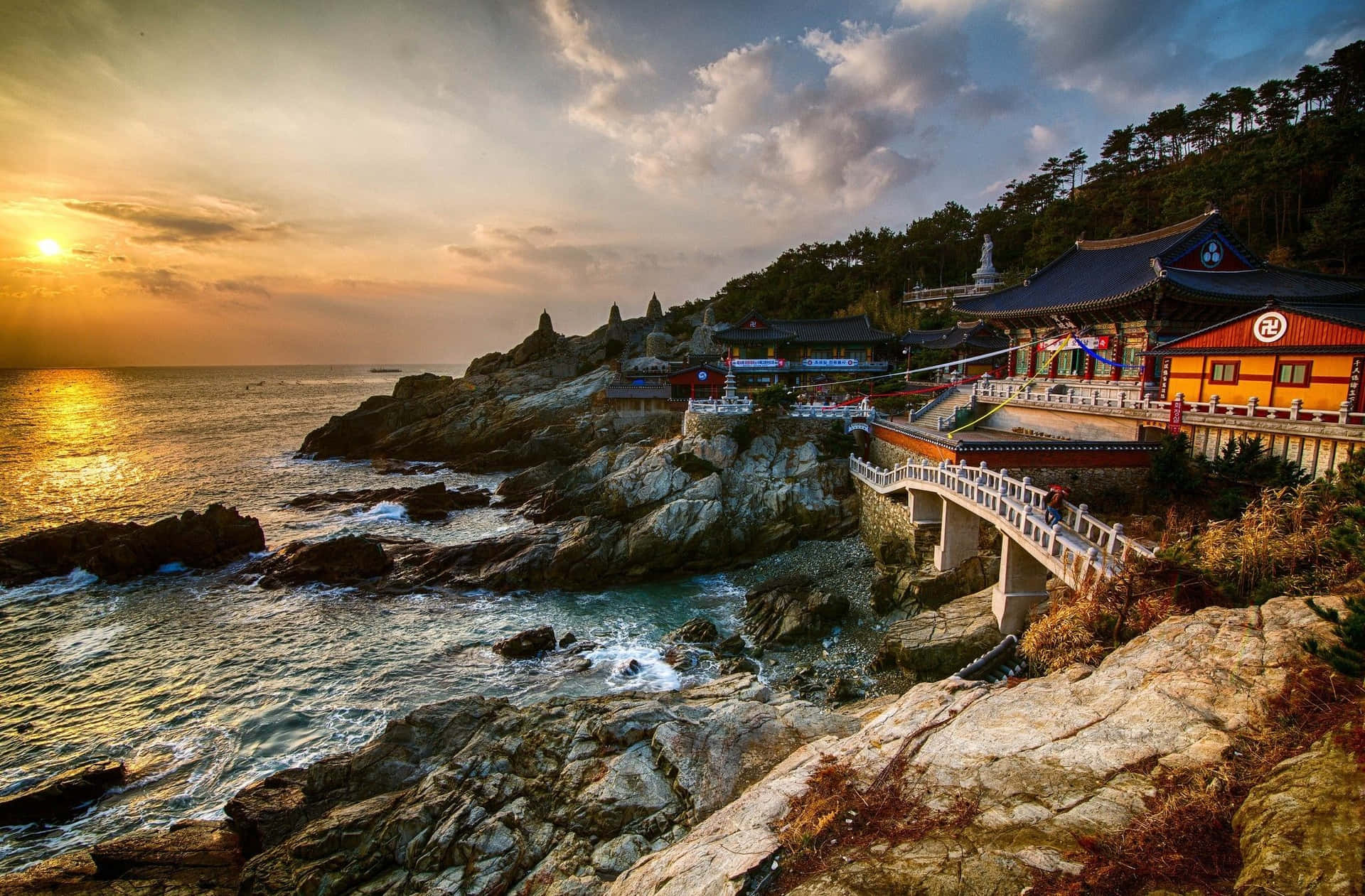 韓國美麗海灘風光 壁紙 — 夕陽灑落在岩石懸崖上的海岸寺廟建築群，下方海浪拍打。