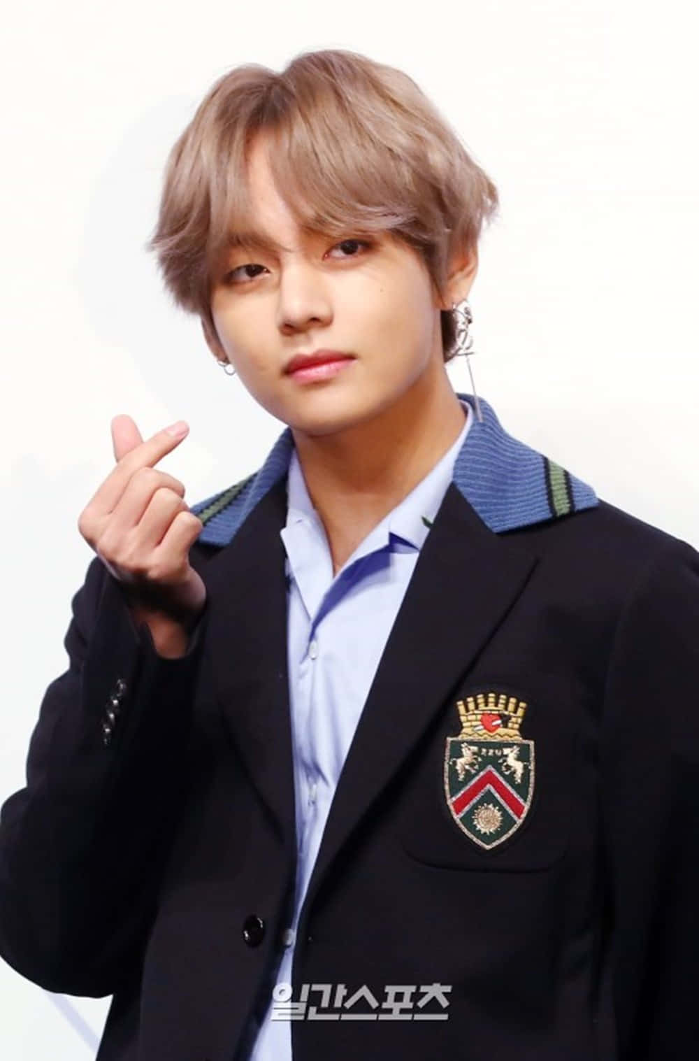 韓國藝人 V BTS 指心 壁紙 — 一位金髮年輕男子穿著帶有徽章的黑色西裝外套，在純白色背景前做手勢。