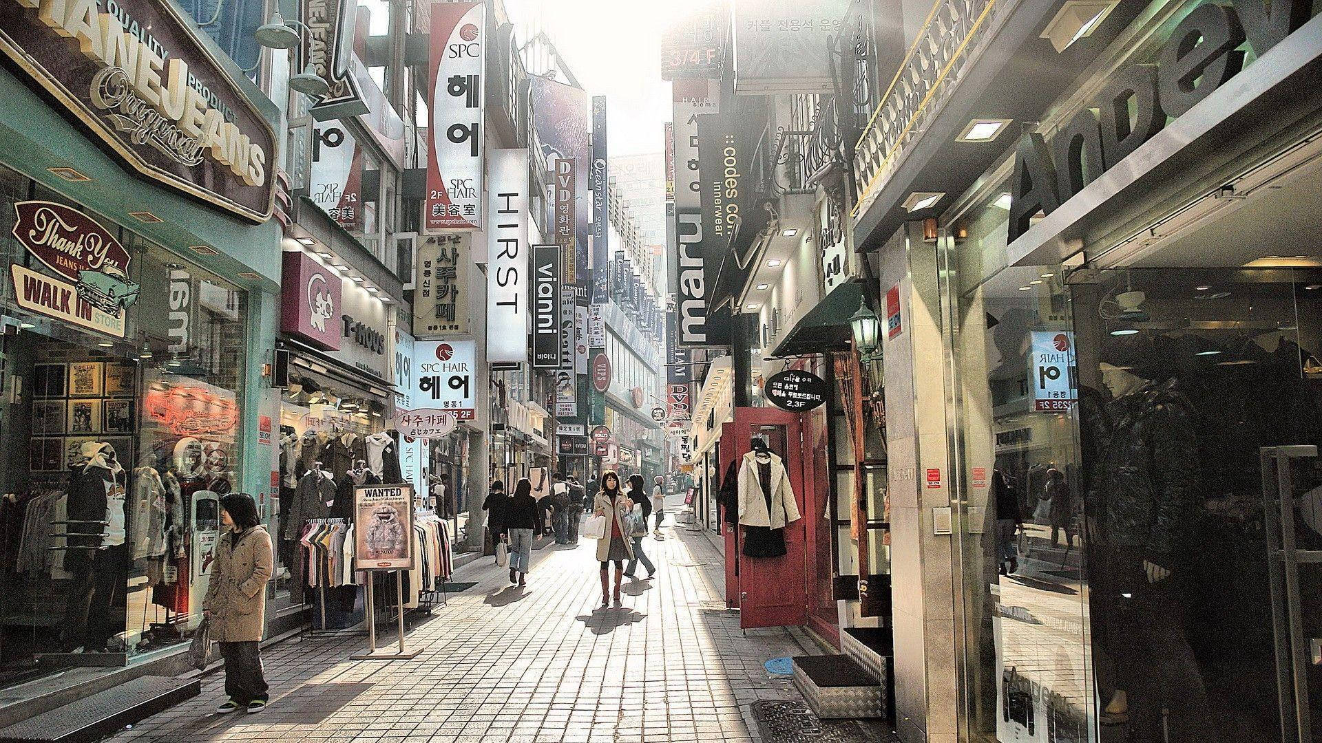 韓國街景 壁紙 — 陽光灑落在一條繁華的城市小巷，兩旁是色彩繽紛的商店門面，行人在其間穿梭。