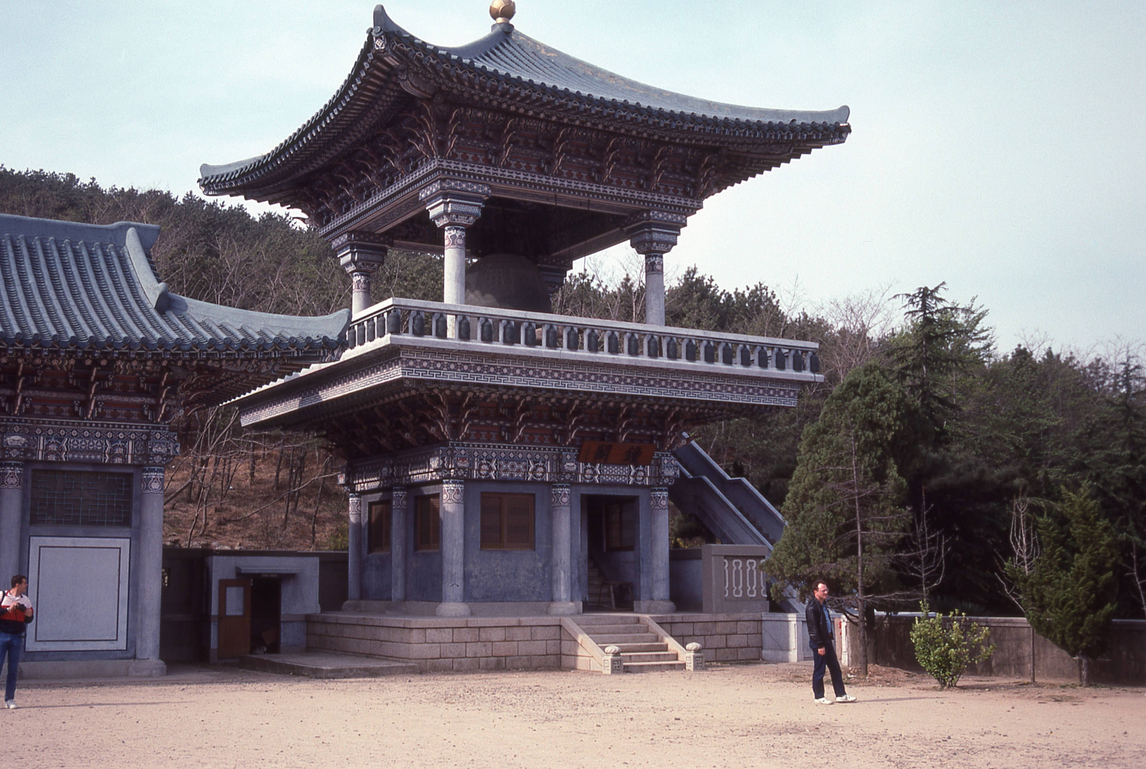 韓國釜山佛寺 壁紙 — 一座傳統東亞寺廟，擁有華麗的瓦頂和石柱，矗立在樹木之間，天空多雲。