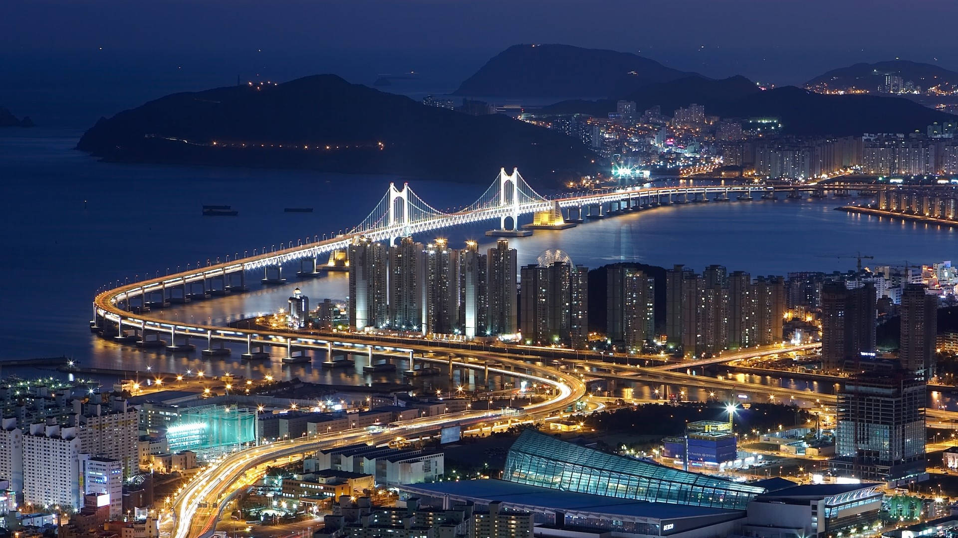 韓國釜山市橋夜景 壁紙 — 一條發光的吊橋橫跨海灣，下方是燈火通明的城市景觀和蜿蜒的高速公路，壯麗的夜景。