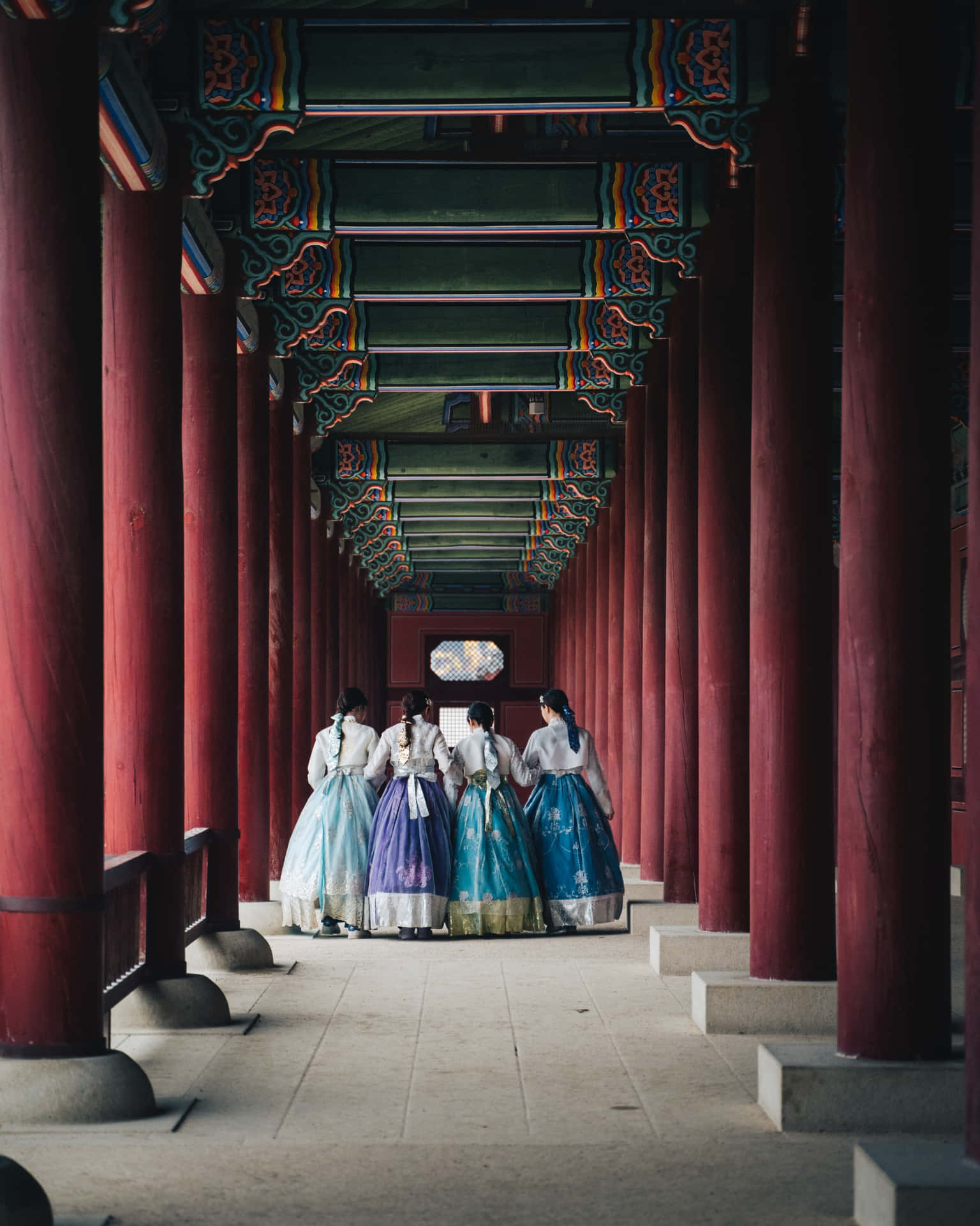 韓國首爾繁忙的城市景觀 壁紙 — 四位身穿色彩繽紛韓服嘅女士，喺由紅色柱同華麗綠色天花框住嘅傳統韓國寺廟走廊行開。