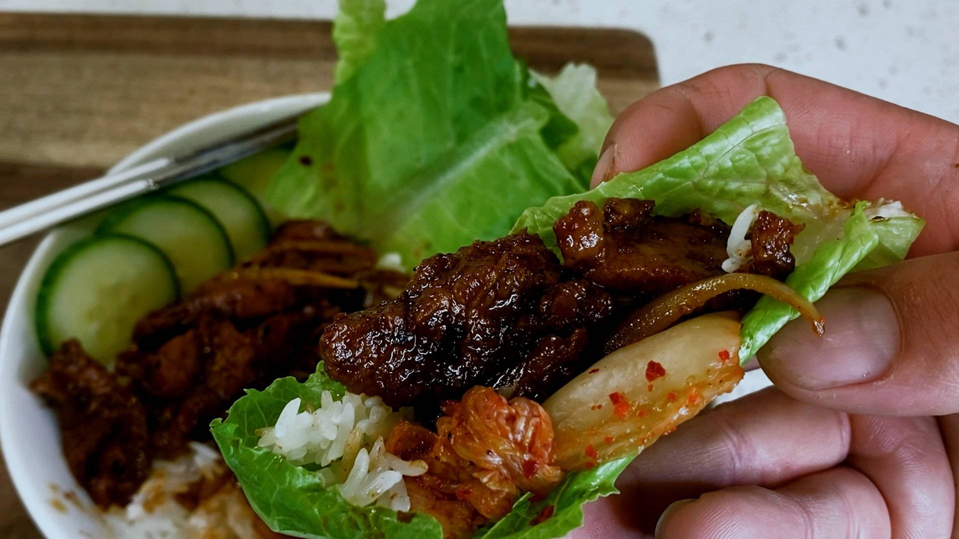 韓式烤肉包生菜 壁紙 — 一隻手拿著一個生菜包，裡面有照燒肉、泡菜和米飯，放在一碗米飯、青瓜和蔬菜上面。