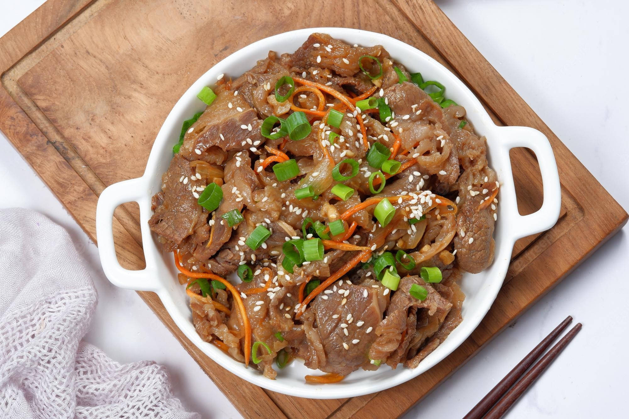 韓式醬油牛肉配胡蘿蔔與蔥 壁紙 — 一個白色陶瓷碗盛載著美味的韓式牛肉烤肉，上面撒有芝麻、青蔥和切絲紅蘿蔔，放在木砧板上，旁邊有筷子。