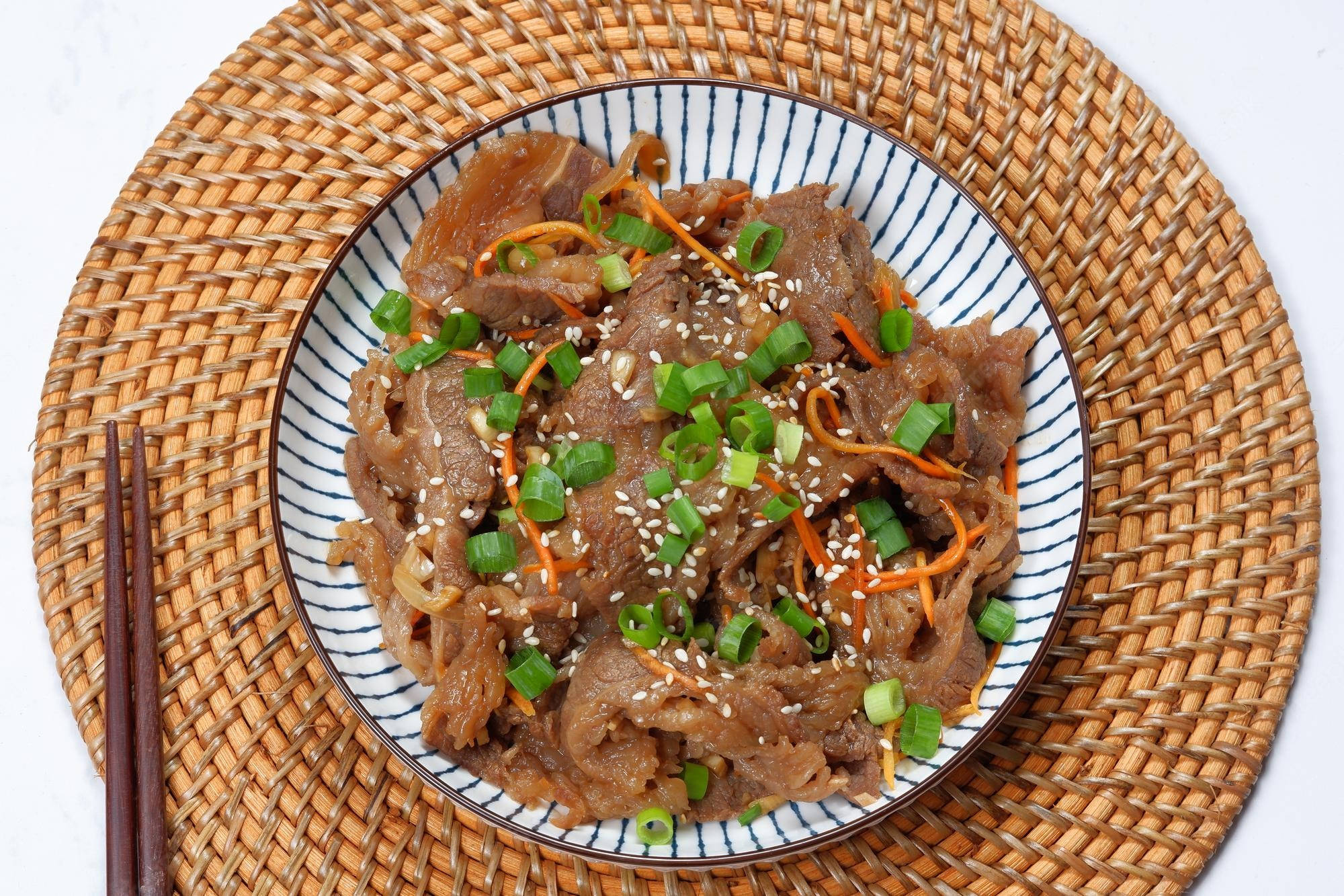 韓式醬油牛肉配芝麻與胡蘿蔔 壁紙 — 一碟美味牛肉，上面撒有芝麻、青蔥和蘿蔔絲，放在編織墊上，旁邊有筷子。