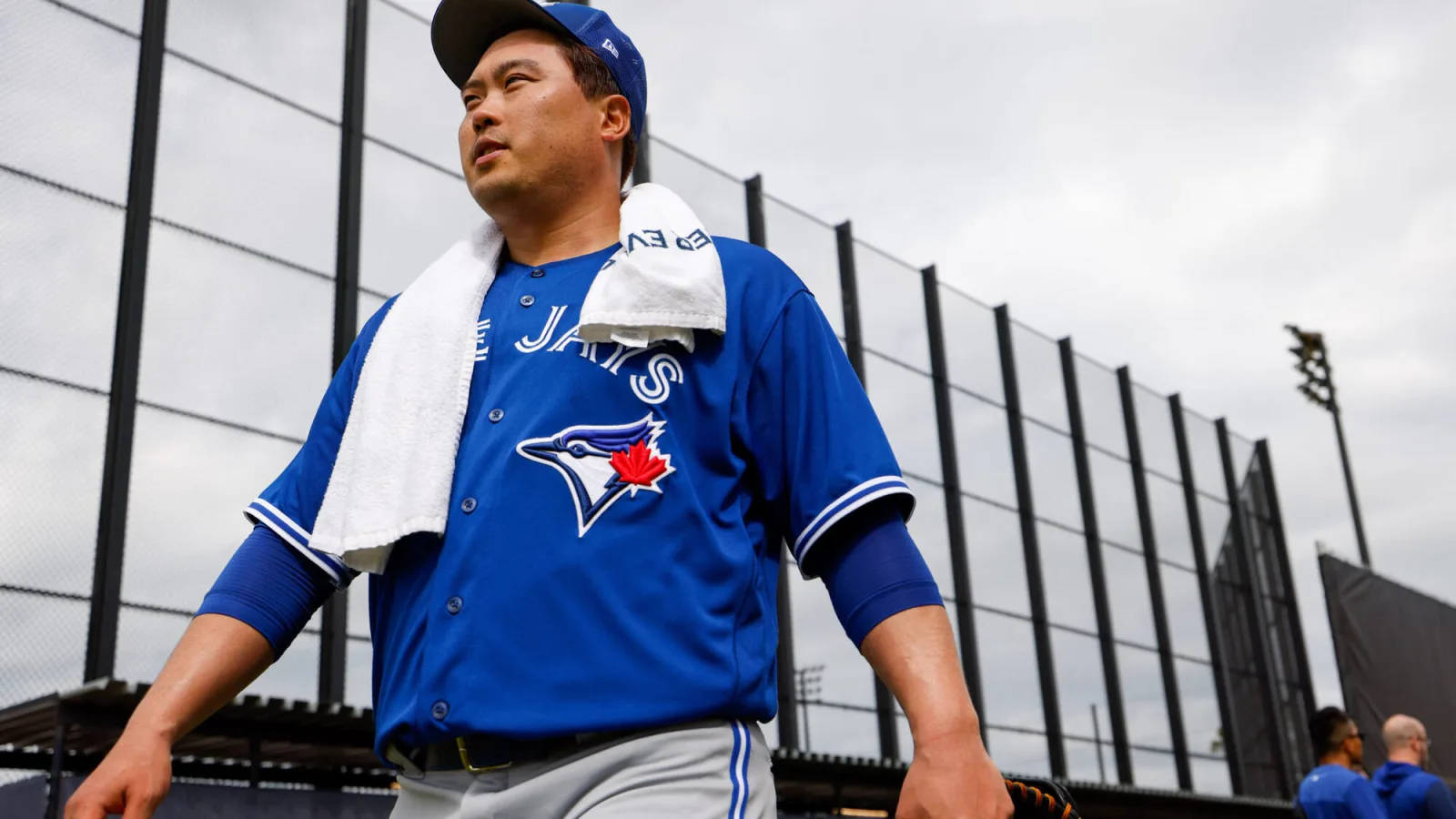 身穿藍鳥隊球衣的棒球運動員，肩上搭著毛巾，走近球場圍欄。