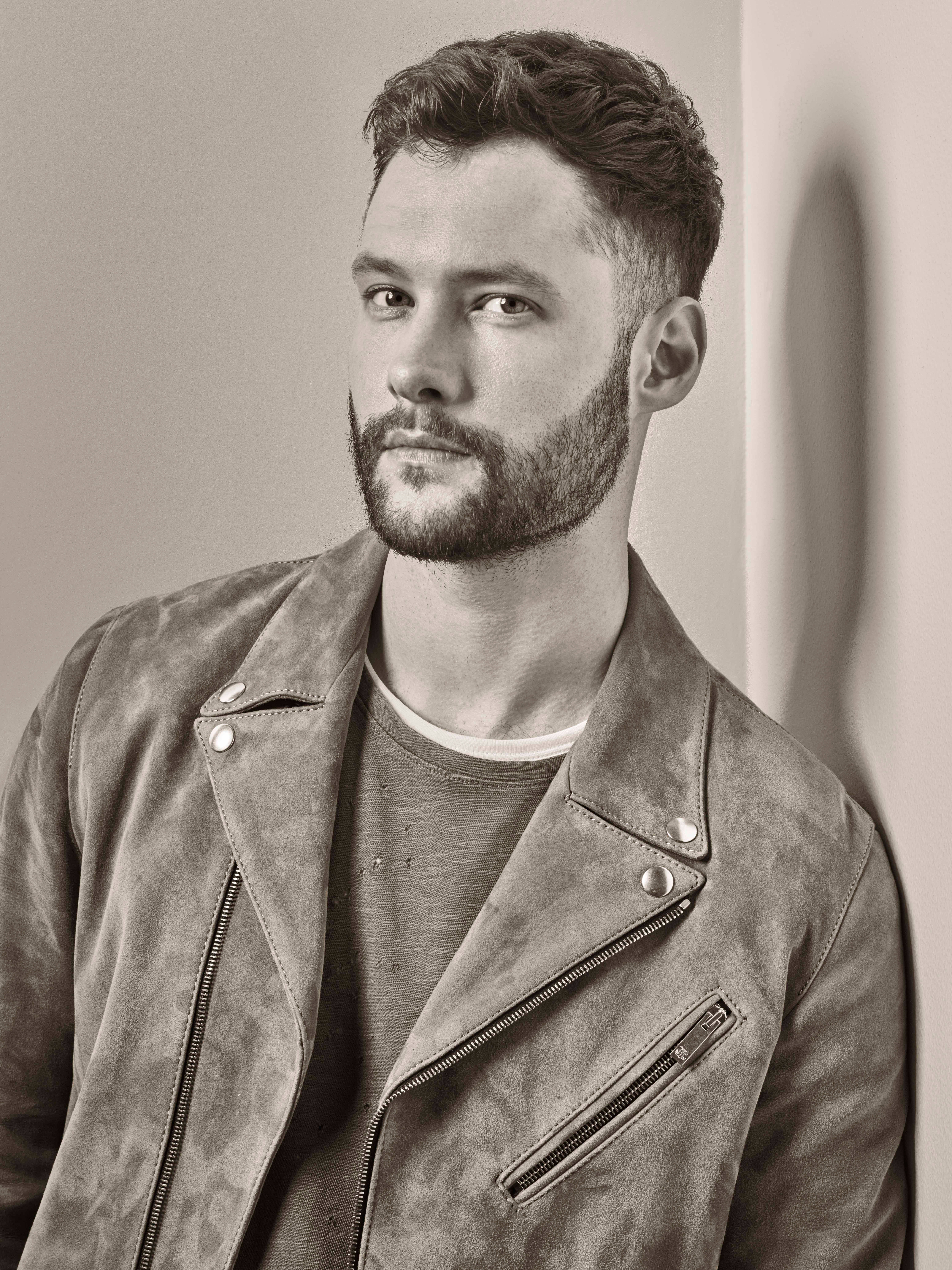 音樂人 Calum Scott 壁紙 — 一位留鬍鬚的英俊男士，穿著質感皮革外套，靠著素色牆壁，呈現憂鬱的黑白肖像。