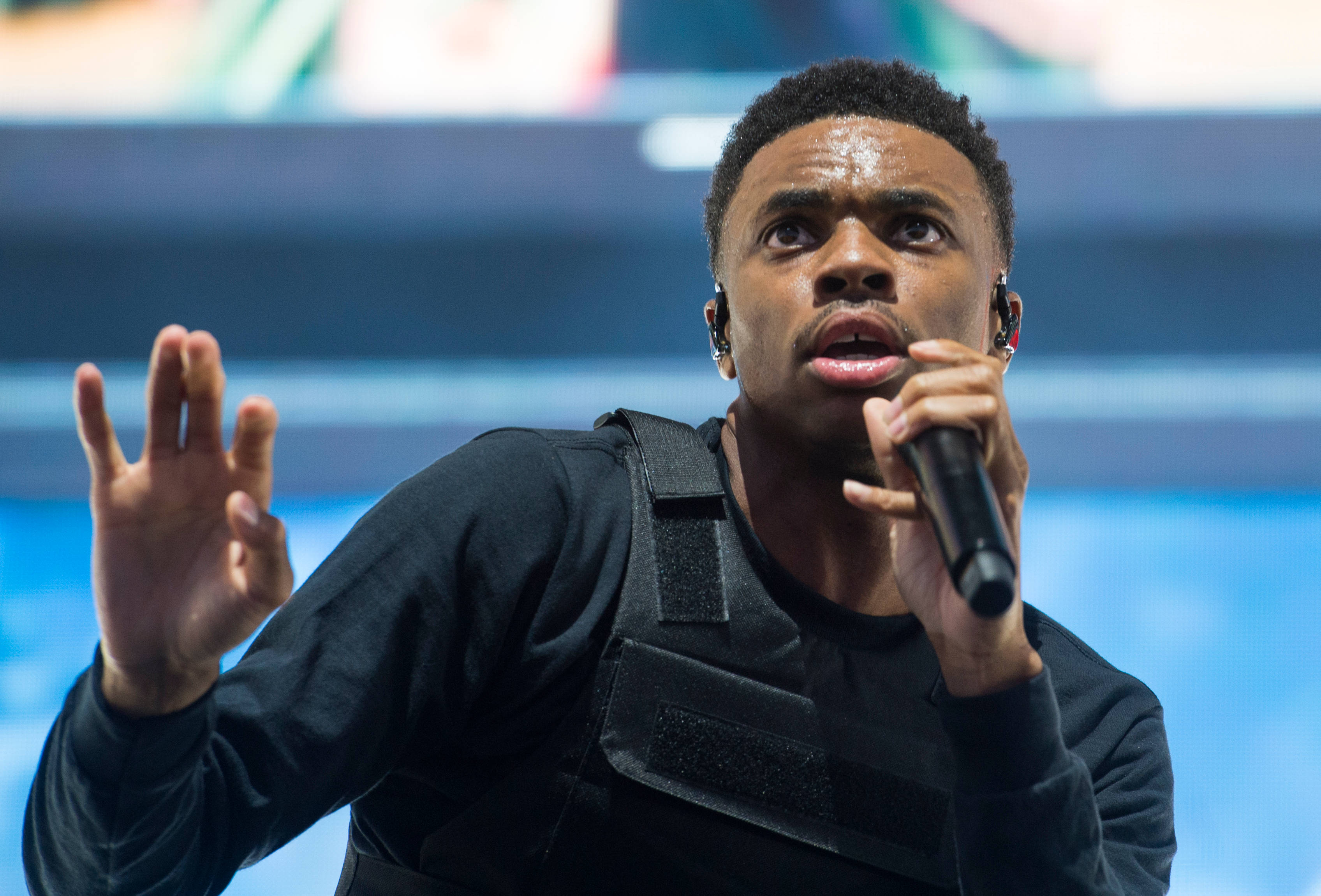 音樂人 Vince Staples 壁紙 — 一位身穿黑色裝備的男表演者在燈光明亮的舞台上，一手舉起，熱情地對著咪高峰唱歌。