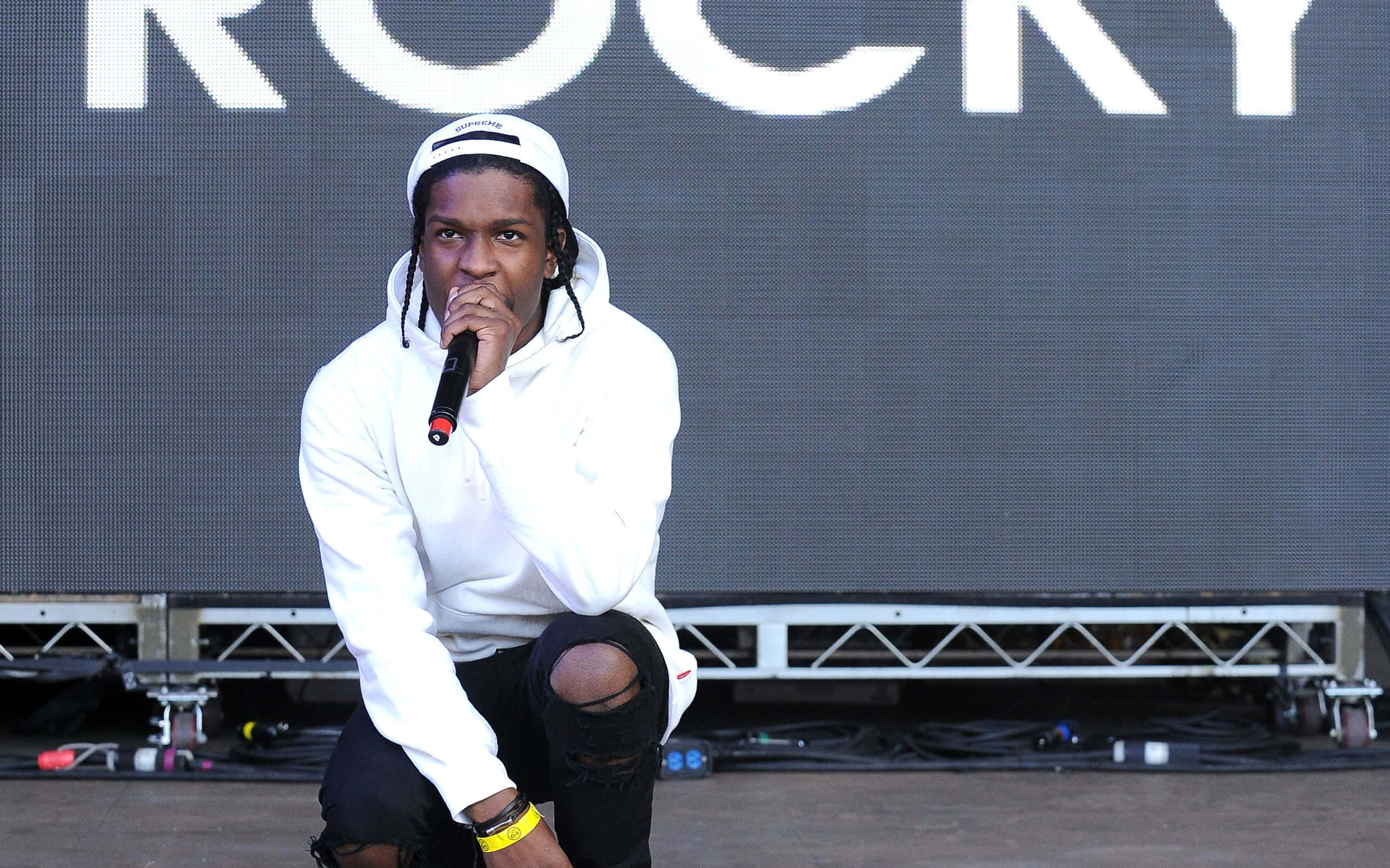 音樂傳奇人物 Asap Rocky 壁紙 — 一位身穿白色連帽衫的饒舌歌手跪在舞台上，在大型「ROCKY」屏幕下拿著麥克風。