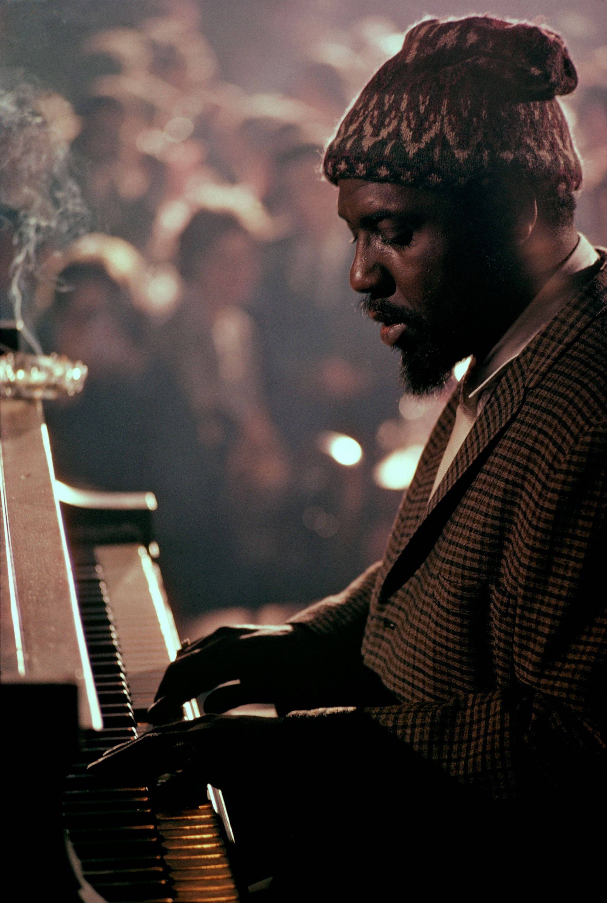 音樂家 Thelonious Monk 壁紙 — 一位戴著針織帽的爵士鋼琴家在溫暖的舞台燈光下熱情演奏，背後是模糊的觀眾。