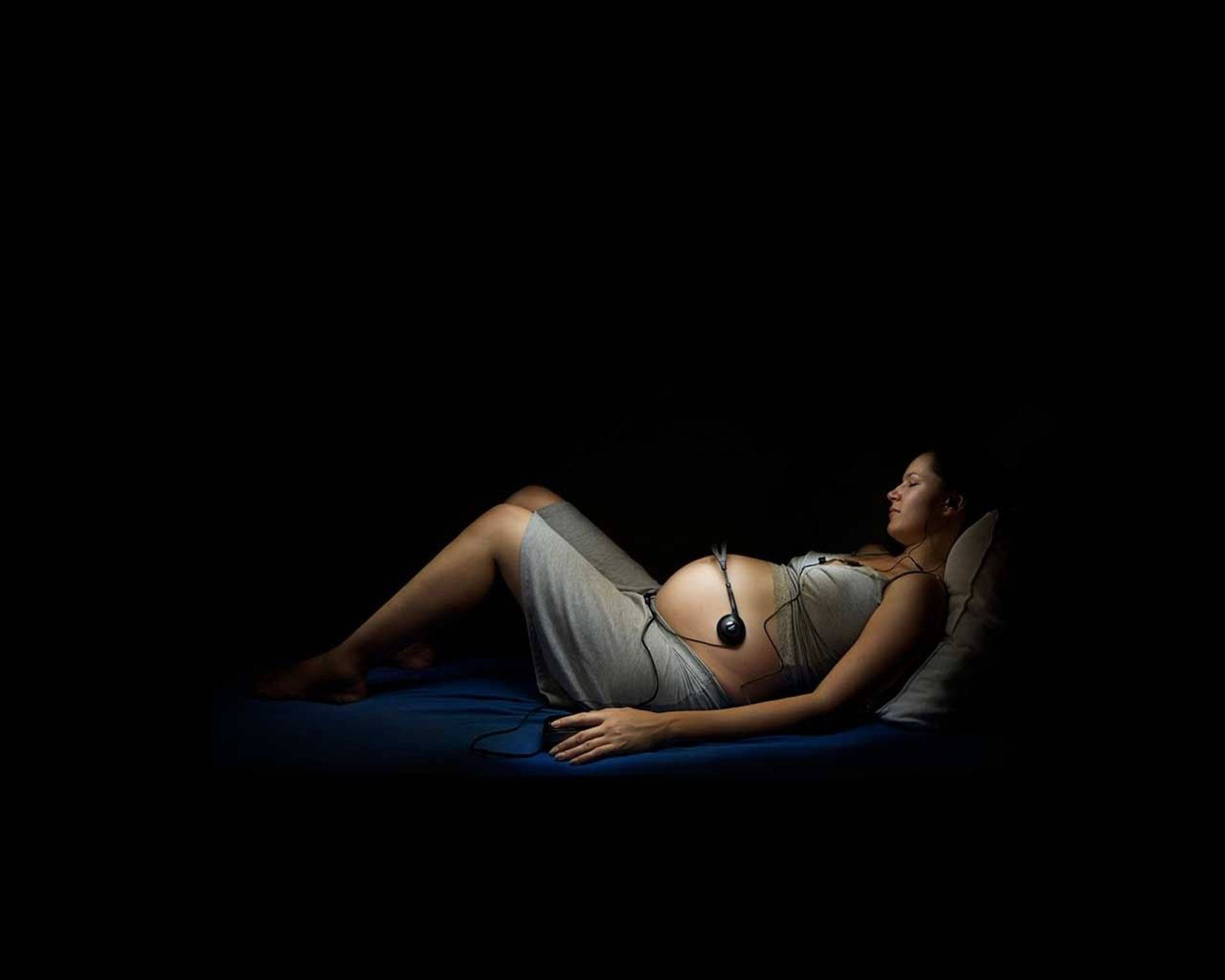 音樂愛好者孕婦 壁紙 — 一名孕婦在黑暗中平靜地躺在藍色表面上，戴著耳機，以平靜的表情向上凝視。