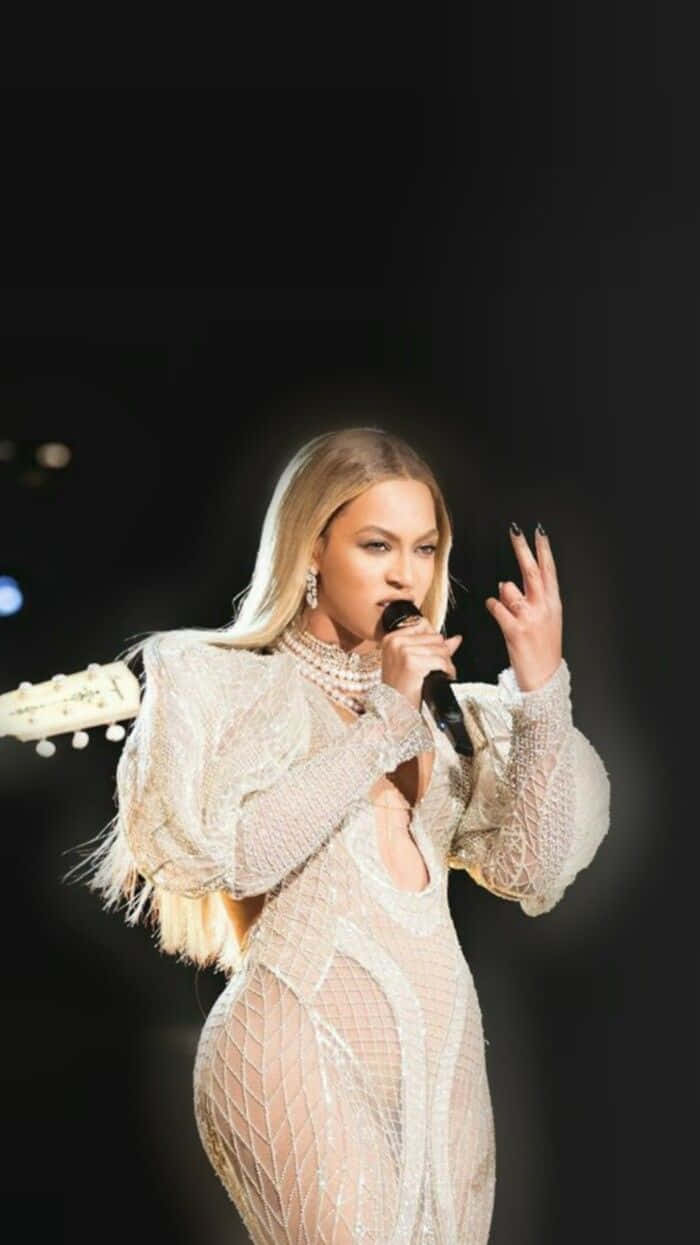 音樂皇後碧昂絲 壁紙 — Beyonce 在舞台上穿著閃閃發光的白色網狀長裙表演，手持麥克風，背後可見結他帶。