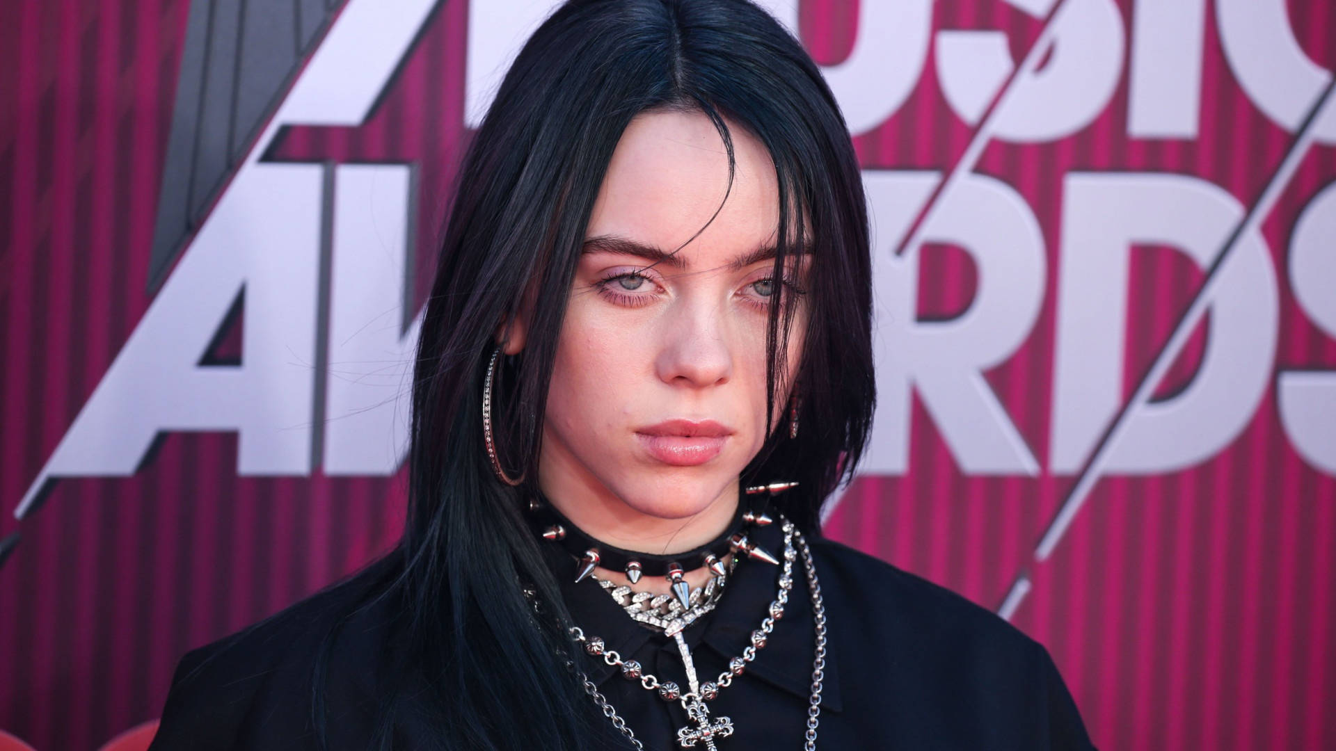 音樂頒獎典禮比莉·艾利什 4K 壁紙 — Billie Eilish 站在紅地毯上，戴著多層鏈條和圈形耳環，背景是鮮明的粉紅色和白色「Music Awards」。