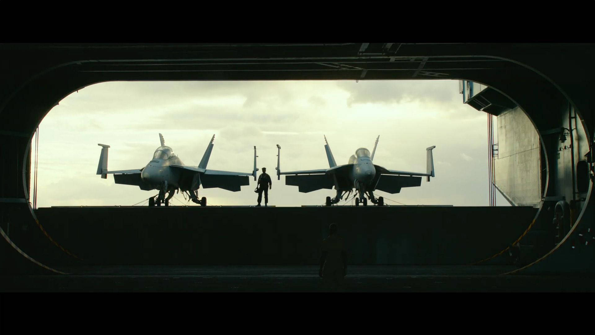 航空母艦甲板上的兩架戰鬥機，一名機組人員在它們之間走過。 — 頂尖飛行員： Maverick 壁紙