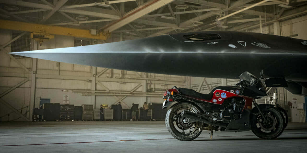 黑色電單車停放在機庫中的隱形戰鬥機下方。 — 頂尖飛行員： Maverick 壁紙