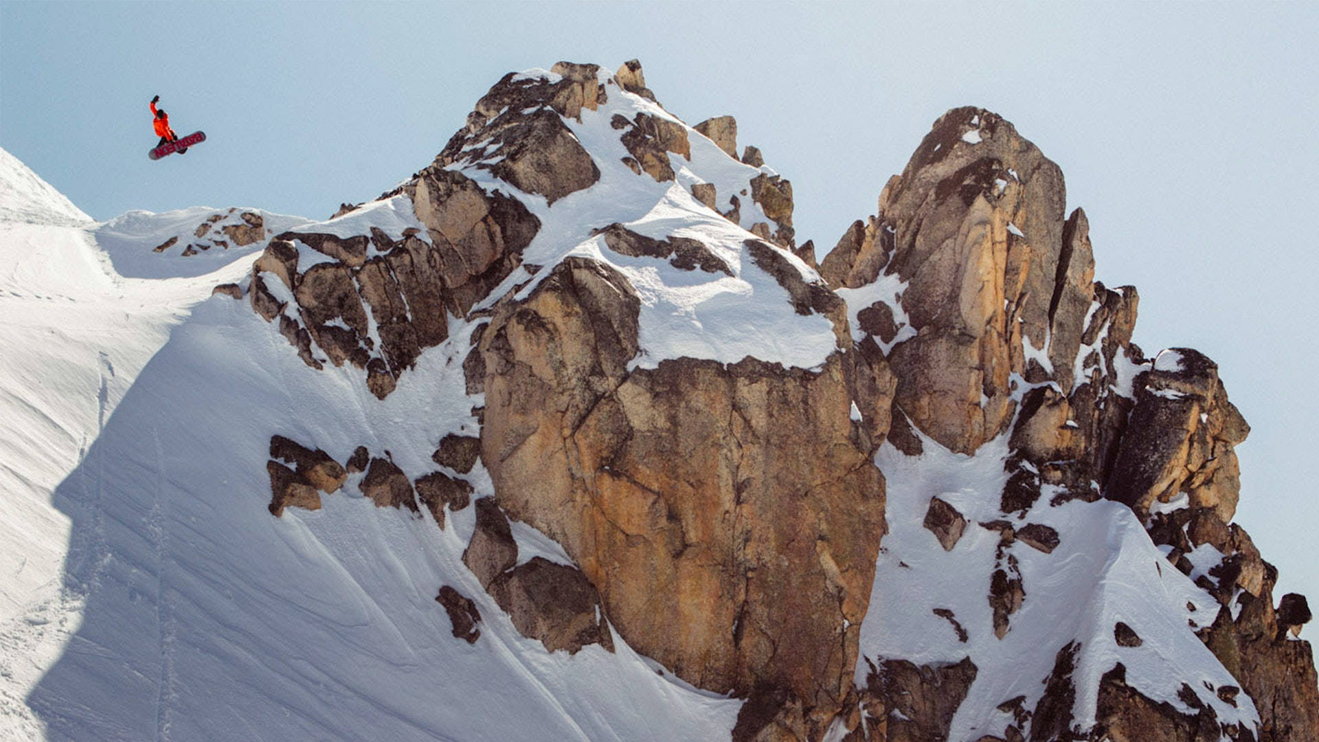 頂峰滑雪 壁紙 — 一名身穿紅色滑雪服的滑雪板運動員在晴朗的藍天下，飛越崎嶇積雪的山峰。