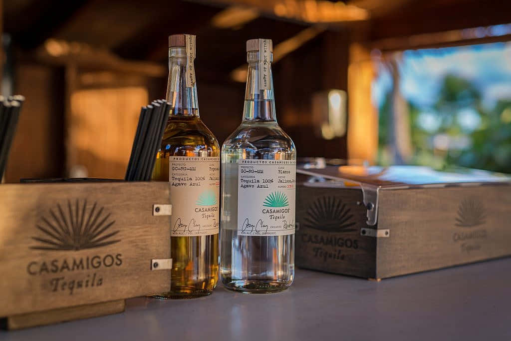 兩瓶Casamigos龍舌蘭酒（琥珀色和透明）放在吧台上，有木箱和熱帶背景。