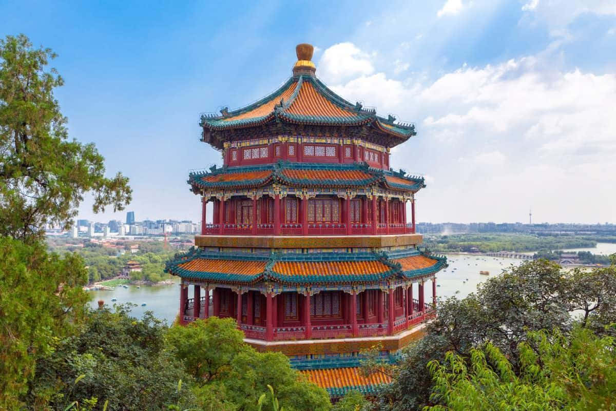 頤和園佛香閣上藍天 壁紙 — 一座色彩鮮豔的紅橙色傳統中國寶塔，配以綠色瓦片屋頂，雄偉地矗立在山坡上，俯瞰著寧靜的湖泊和遠處的城市景觀，在明亮的藍天下。