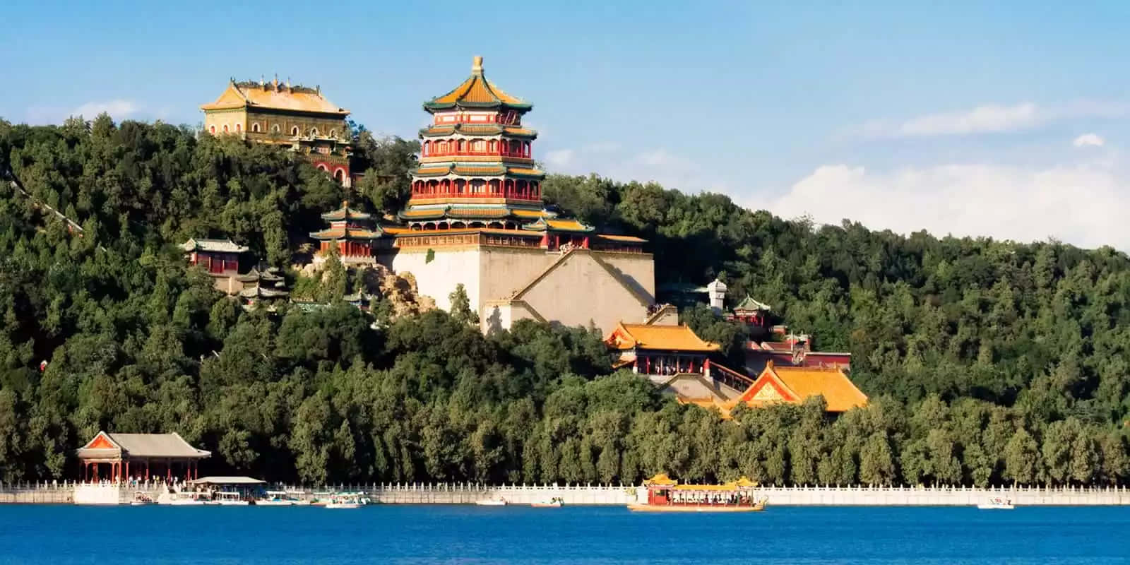 頤和園周圍的森林 壁紙 — 宏偉的傳統中國宮殿建築，金色屋頂矗立在鬱鬱蔥蔥的綠色山丘上，俯瞰著寧靜的藍色湖泊，湖上有船。