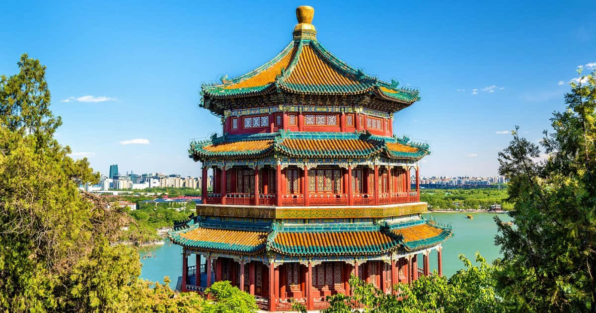 頤和園白天的三層寶塔 壁紙 — 一座色彩鮮豔的紅金色傳統中國寶塔，雄偉地矗立在寧靜的湖邊，周圍是茂密的綠樹，天空清澈湛藍。