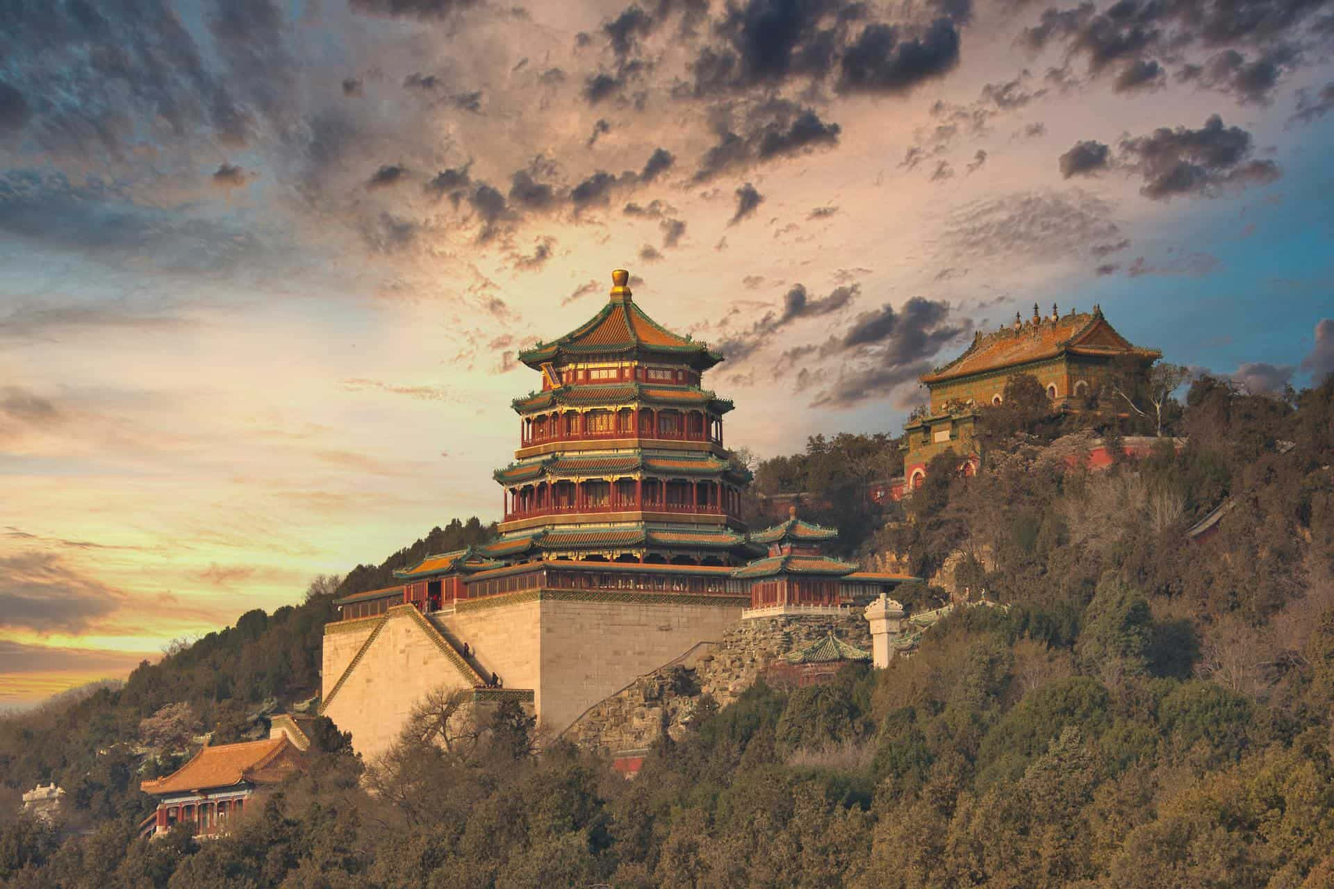 頤和園萬壽山日落 壁紙 — 一座雄偉的傳統中式寶塔，坐落在森林覆蓋的山丘上，背景是壯麗的日落天空。