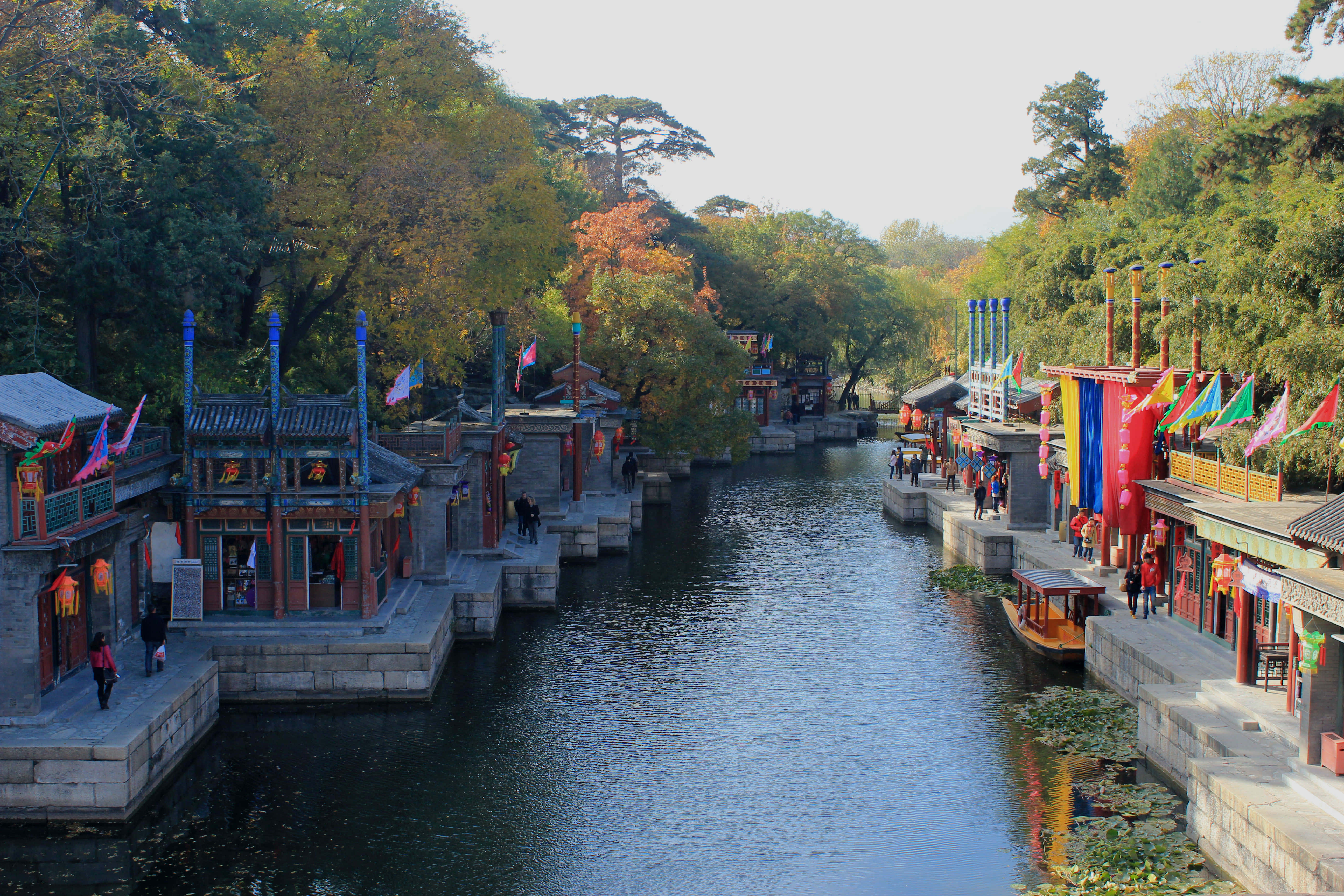頤和園蘇州街市景 壁紙 — 一條寧靜的運河蜿蜒穿過傳統中式建築，兩旁有秋樹和彩色旗幟點綴水道。