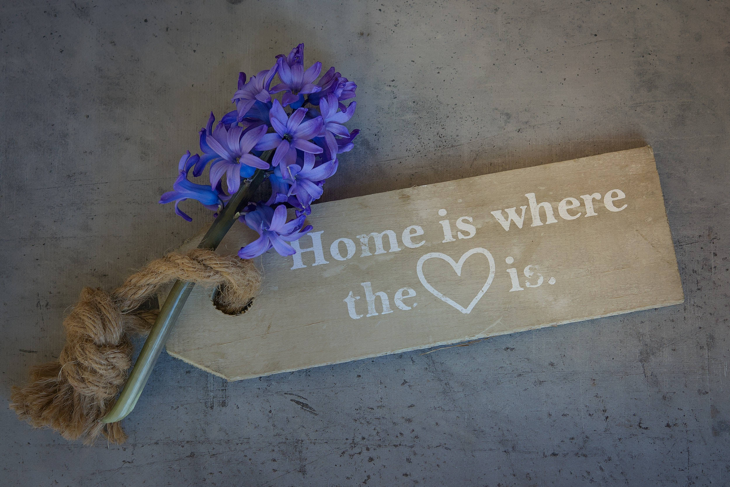 風信子家居牌匾 壁紙 — 一塊寫有「Home is where the ❤️ is」的質樸木牌，放在混凝土表面上，旁邊是一束用麻繩綁著的紫色風信子。